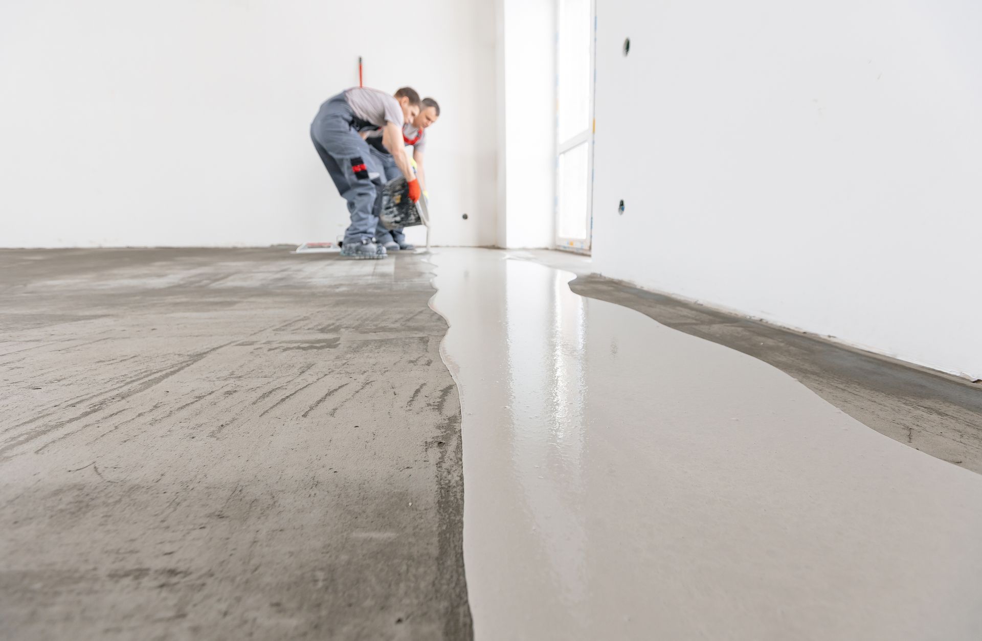 Two workers smoothing concrete floor in a room with white walls, window, and partially smoothed floor.