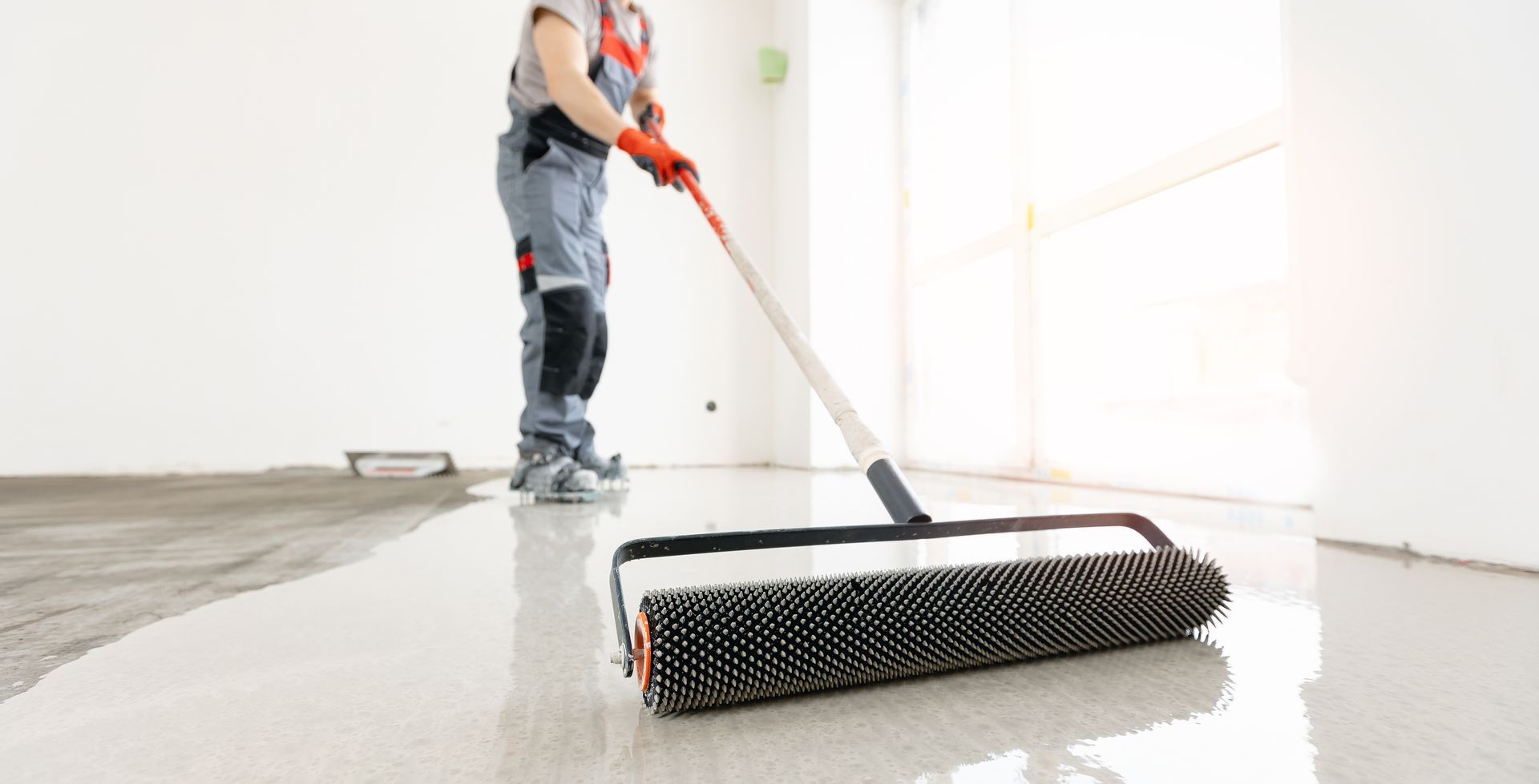 Person rolls a spiked roller over a wet, light-colored floor, likely spreading epoxy; a white-walled room.