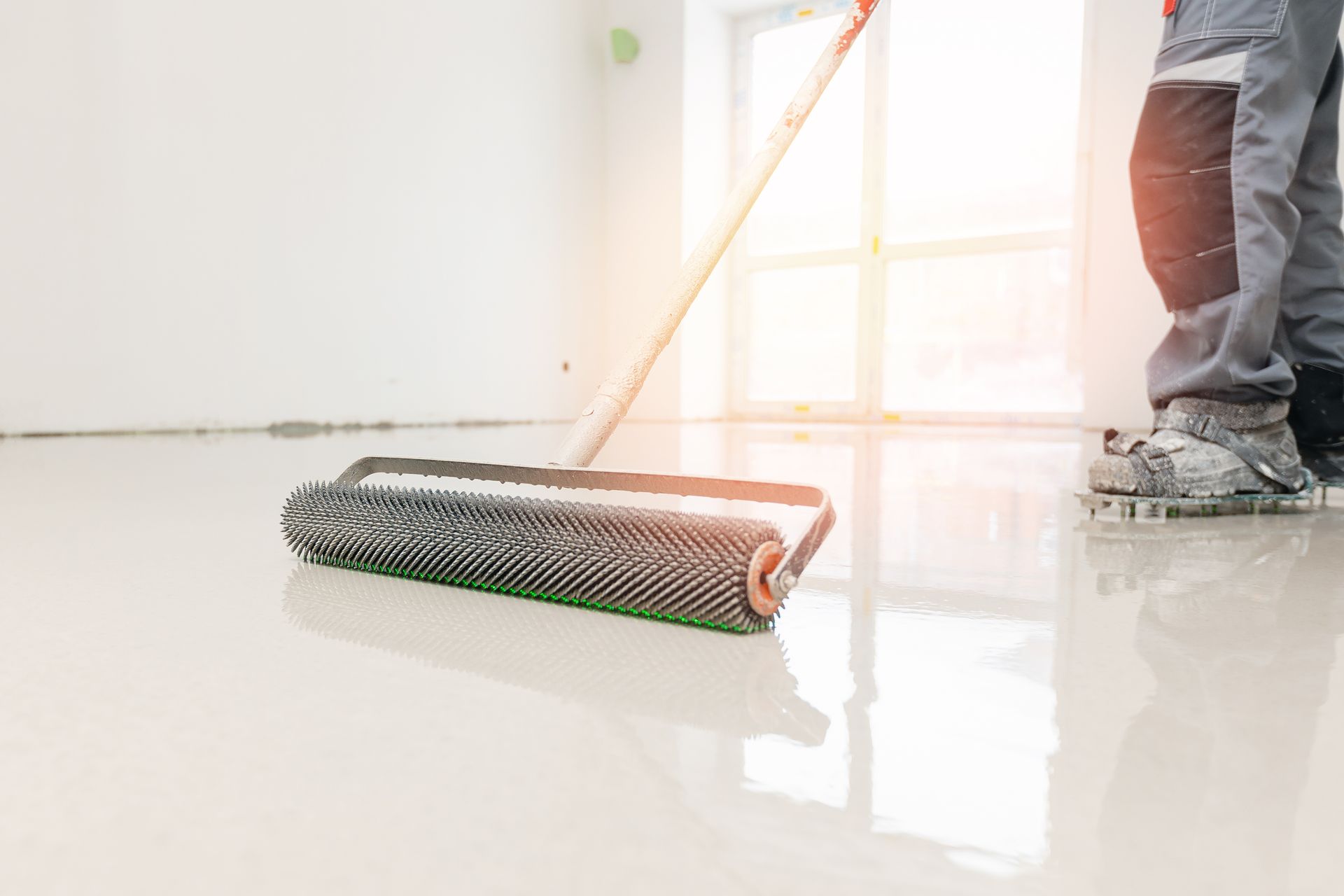 Worker smoothing a light-colored epoxy floor with a spiked roller. Bright light shines through a window.