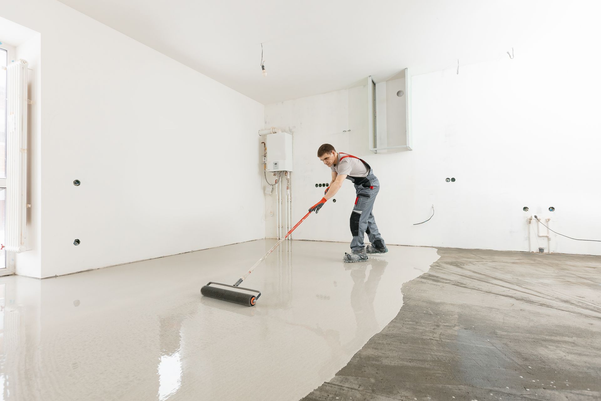 Man applying light gray self-leveling floor compound with a roller in a white-walled room.