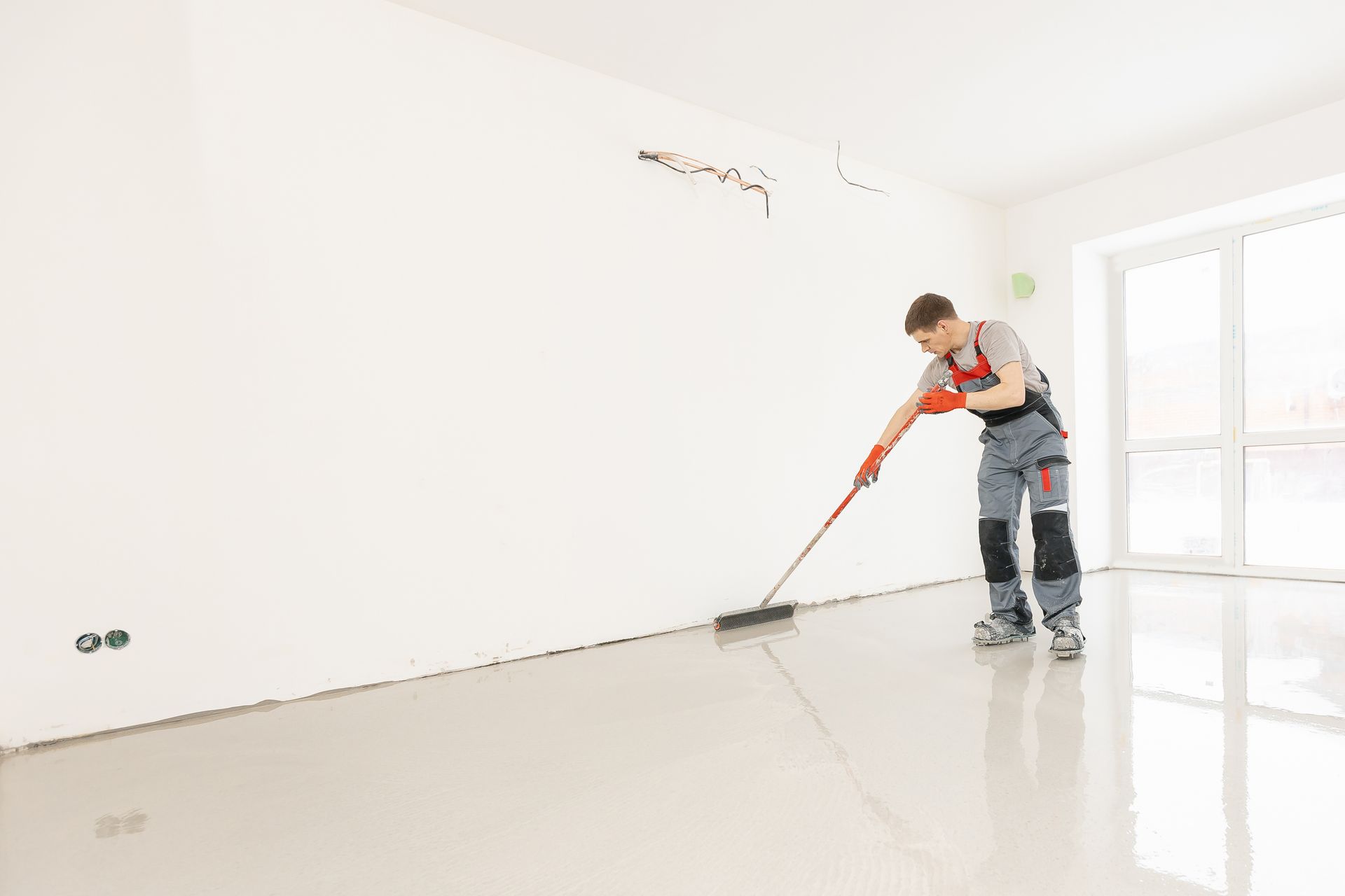 Man smoothing a gray floor with a long-handled tool in a white room with a window.