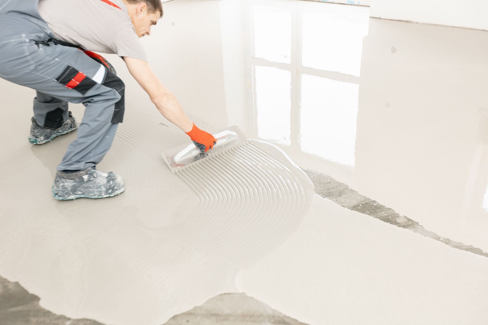 Person applying a self-leveling concrete floor. Wearing safety gear. Interior setting, with window.