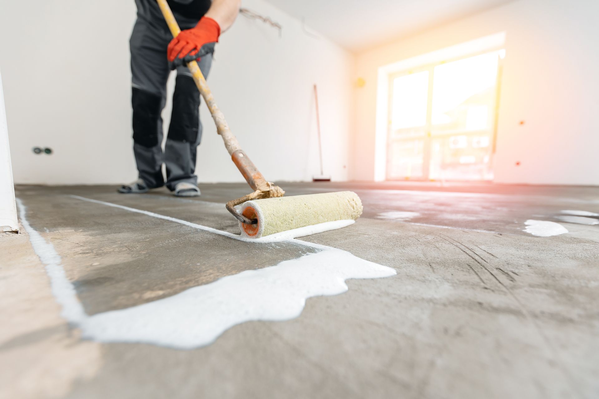 Person in gray pants, orange gloves rolls white paint on concrete floor.