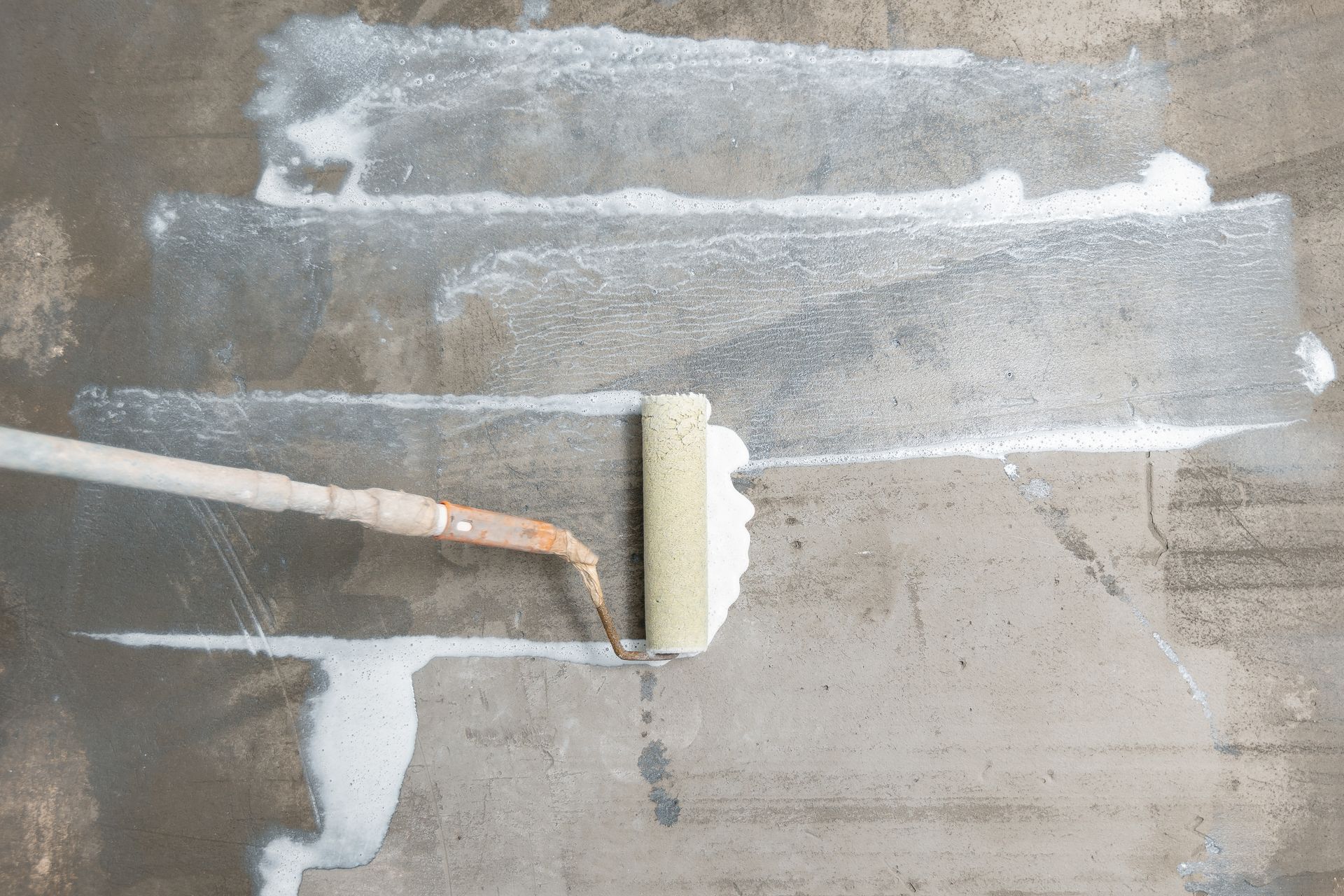 A paint roller applying a white coating to a gray concrete surface.