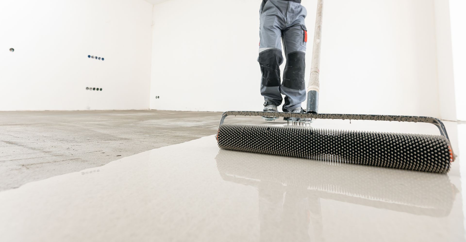 Person using a spiked roller on a freshly poured, light-colored floor, smoothing the surface.