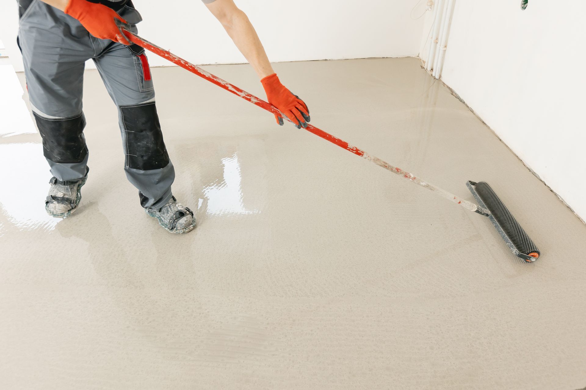 Person in work clothes using a roller to apply a light-colored coating to a floor.
