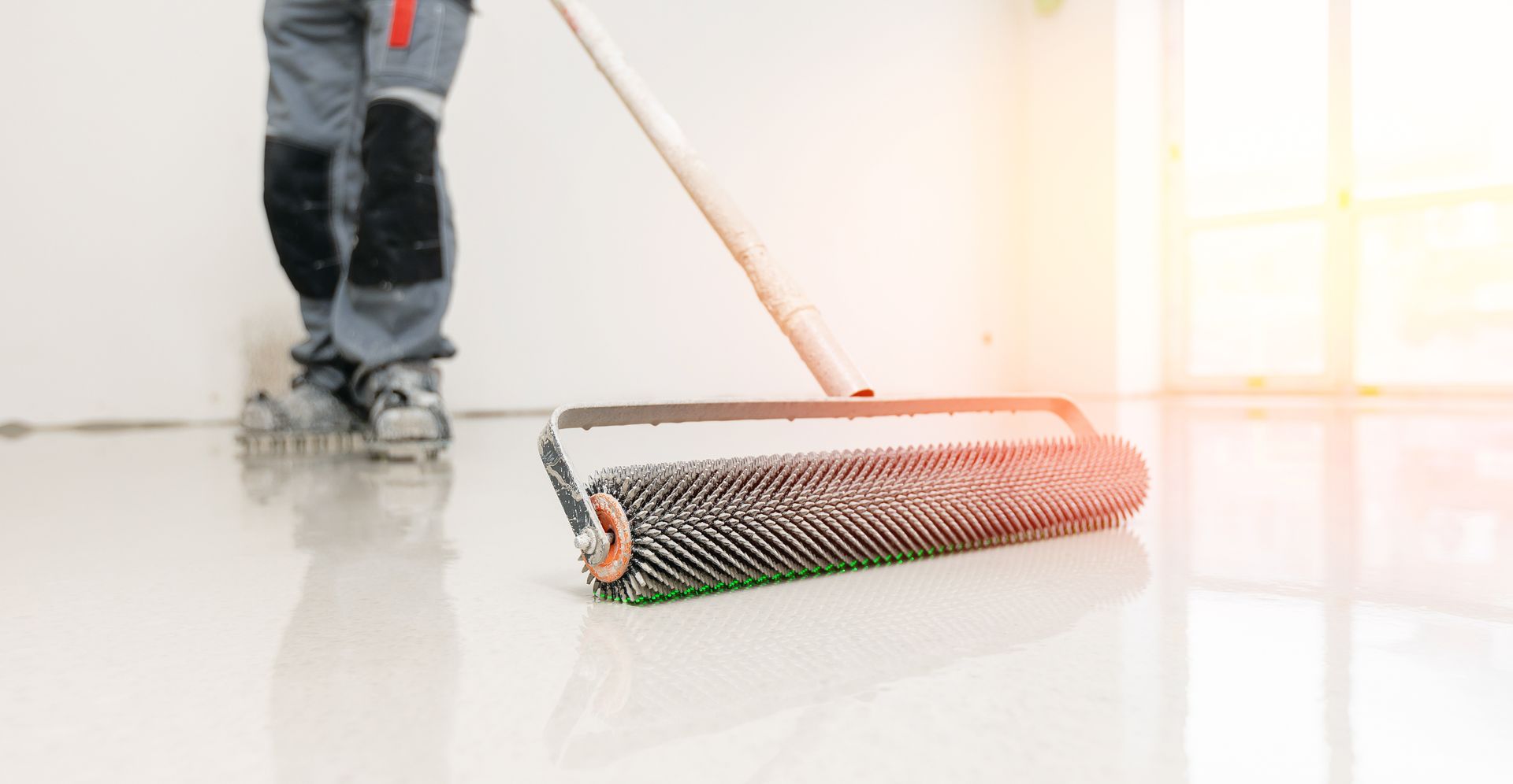 Person applying a glossy coating to a white floor with a spiked roller. Bright interior.