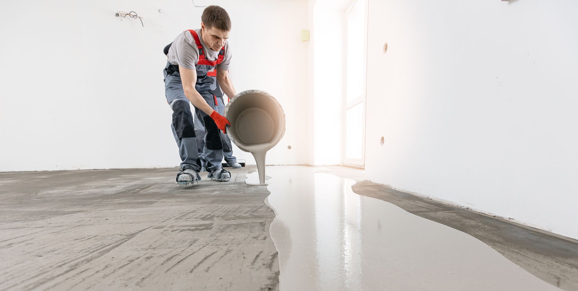 Person pouring concrete from a bucket onto a floor. Indoors, construction site, light gray concrete.
