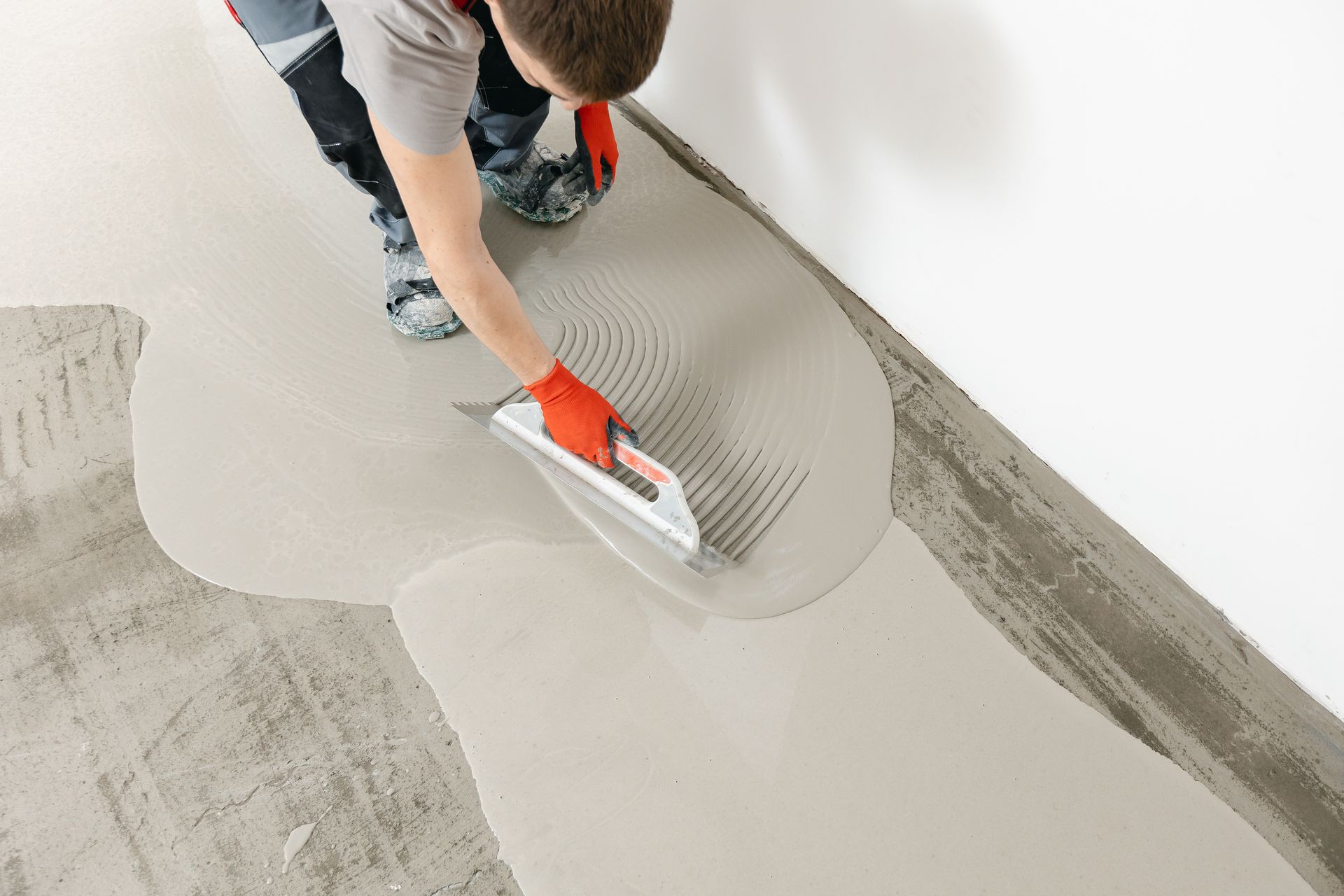 Person applying mortar to a floor with a notched trowel near a wall.