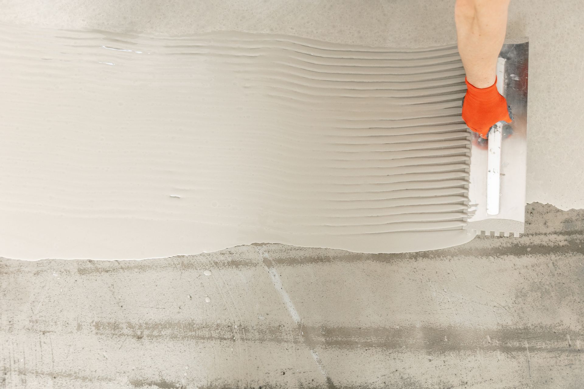 Person in orange glove uses a notched trowel to spread gray adhesive on a concrete floor.