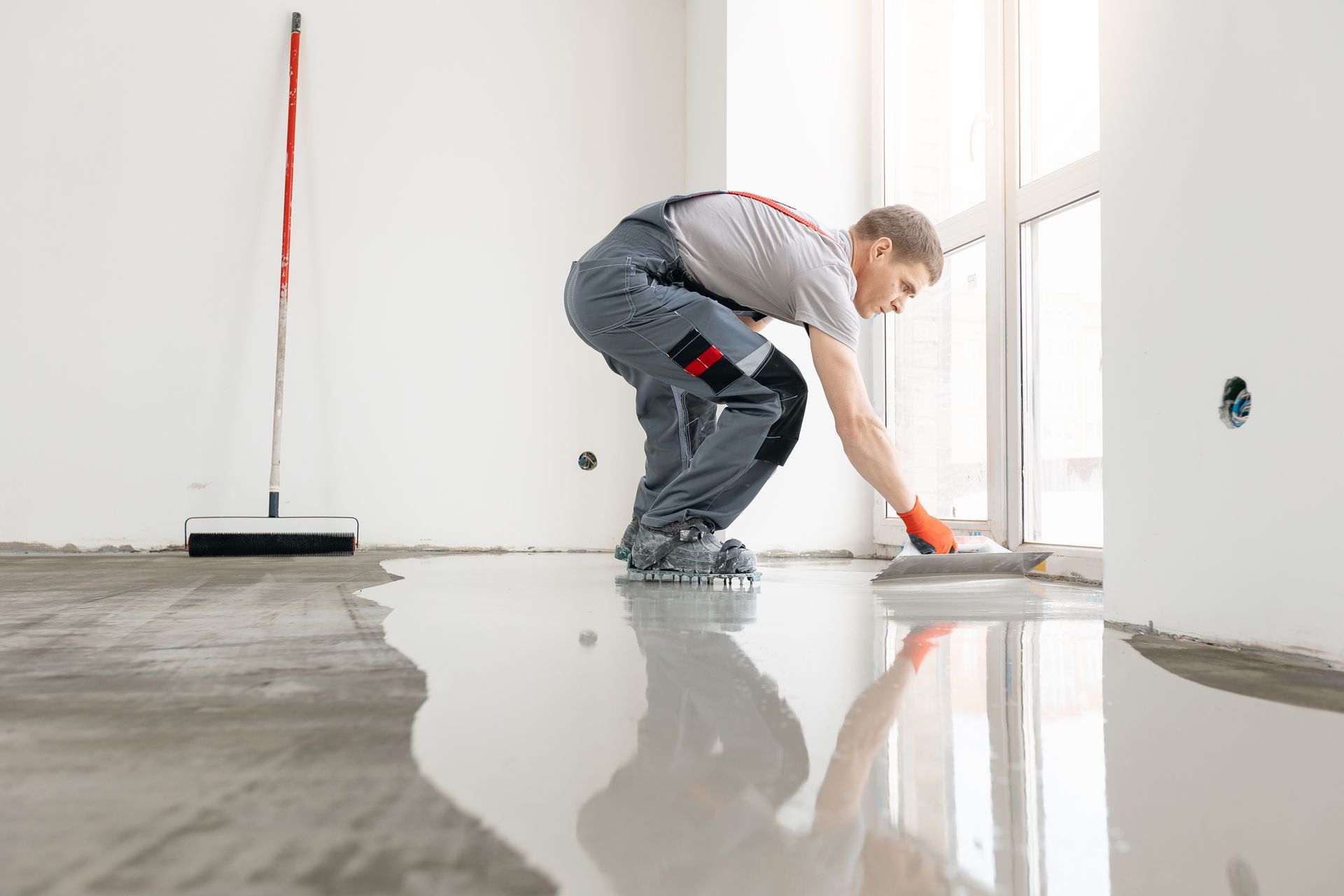 Man leveling a wet, gray floor with a trowel near a window; red-handled tool leans against wall.