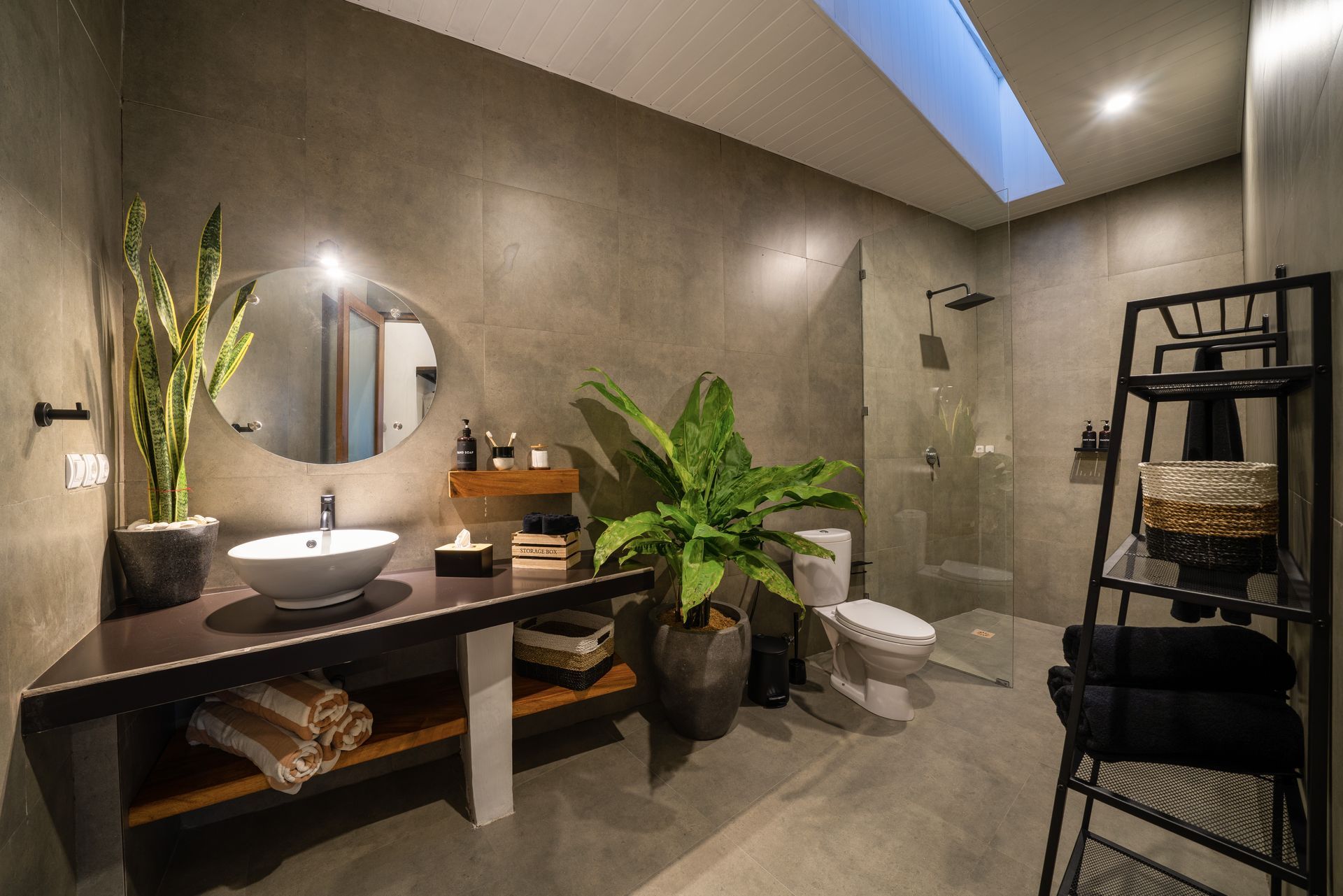 Modern bathroom with concrete walls, oval mirror, plants, and black metal shelving.
