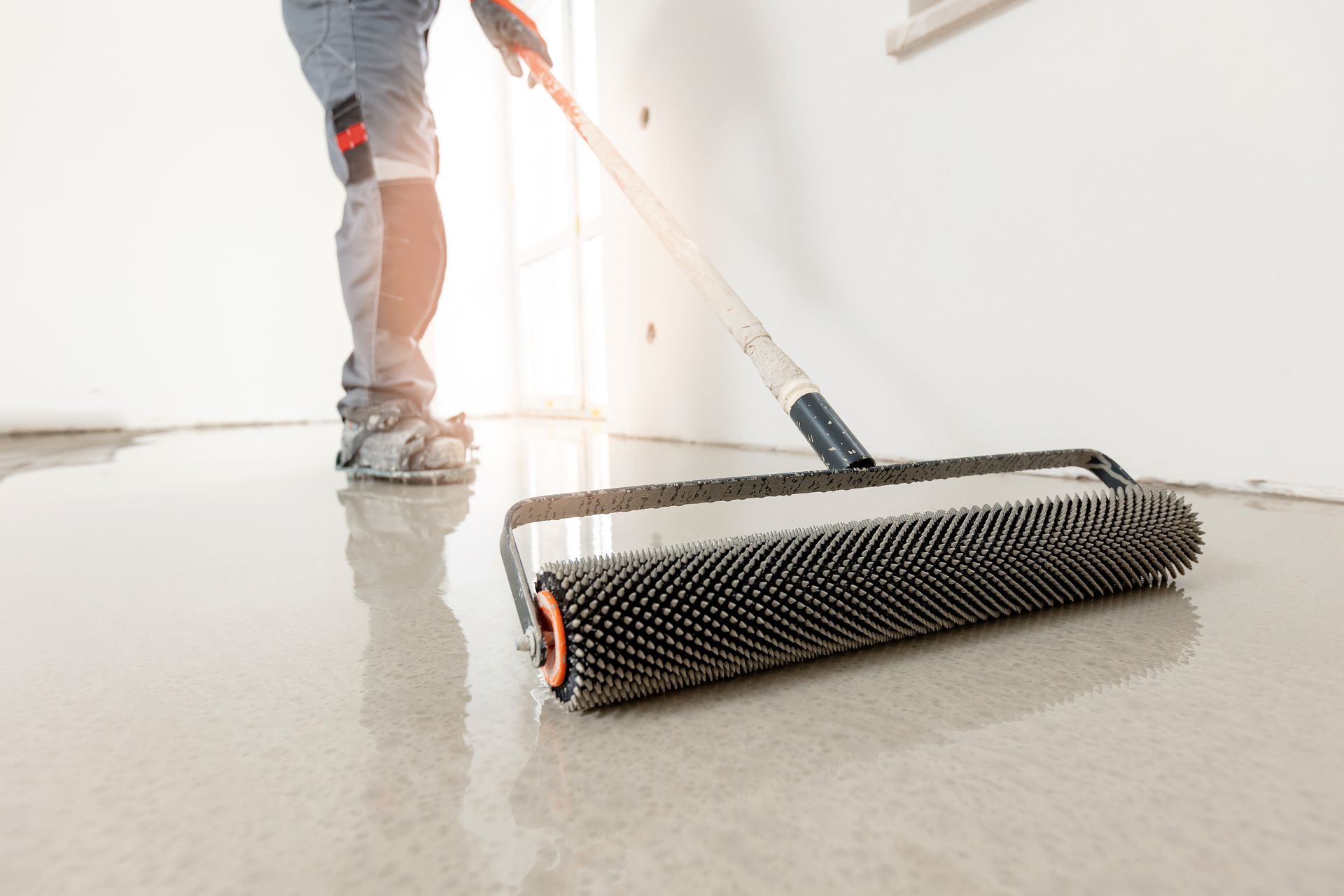 Person smoothing a wet concrete floor with a spiked roller in an indoor setting.