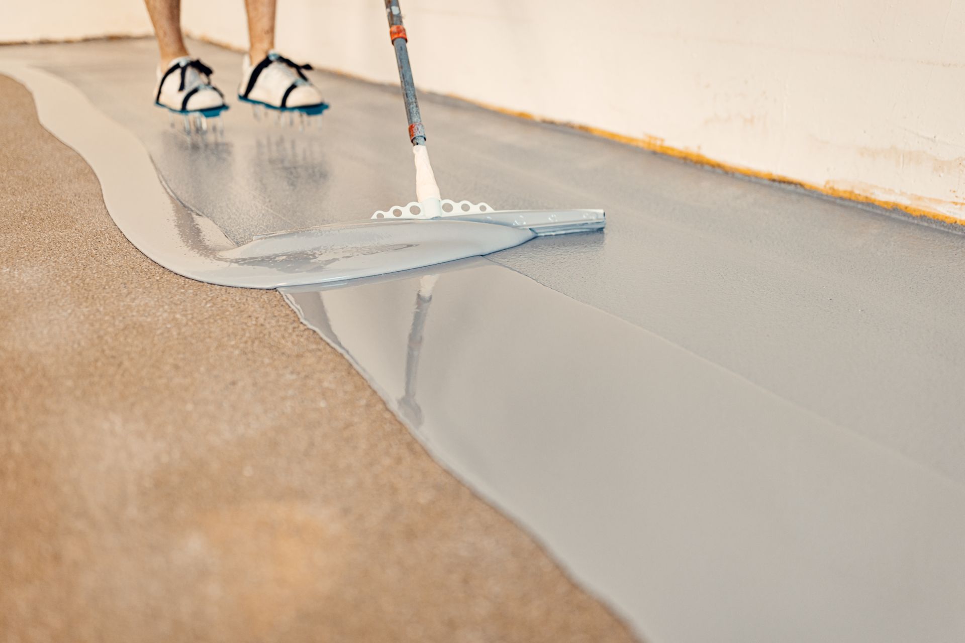 Person applying gray epoxy floor coating with a mop on a concrete surface.