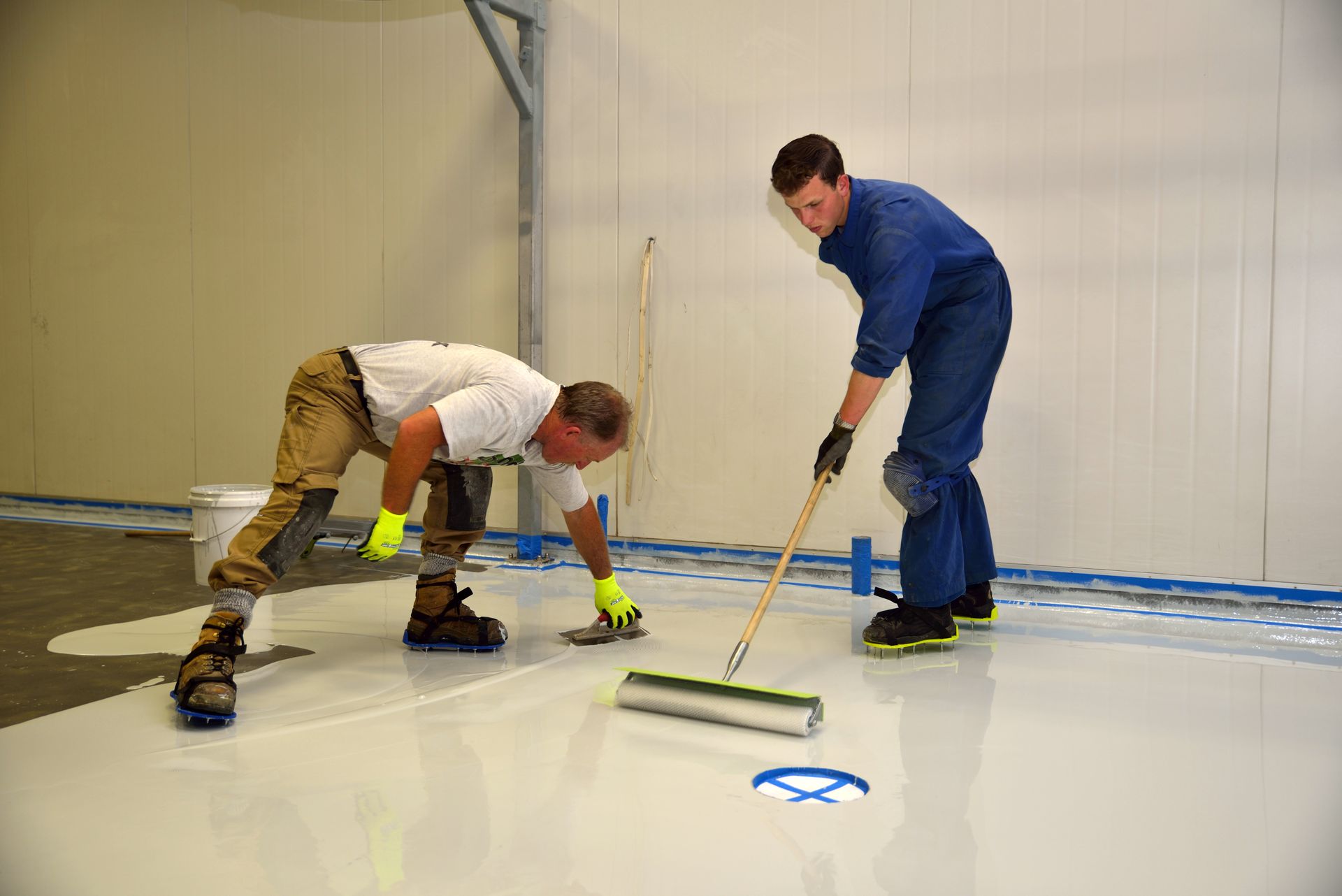 Two workers apply epoxy floor coating in a large, white-walled room, one spreading, the other rolling.