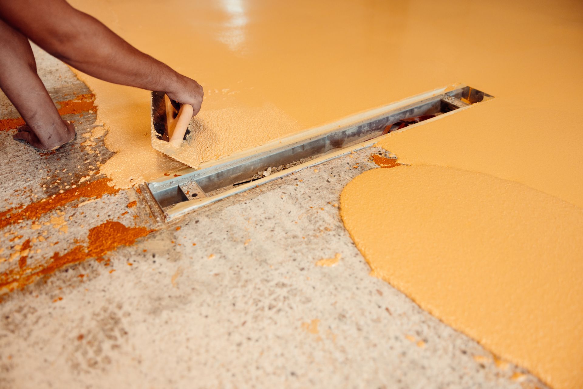 Person smoothing orange epoxy flooring with a trowel, near a drain.