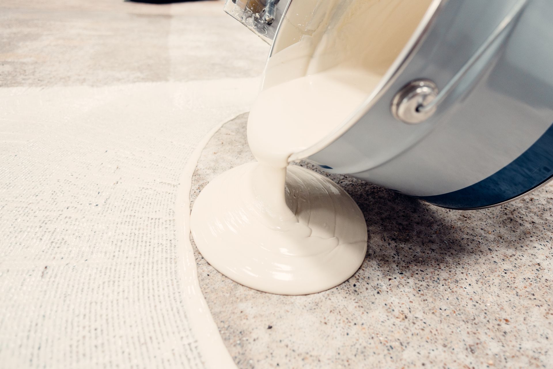 Creamy white substance being poured from a silver bucket onto a textured surface, possibly for a project.