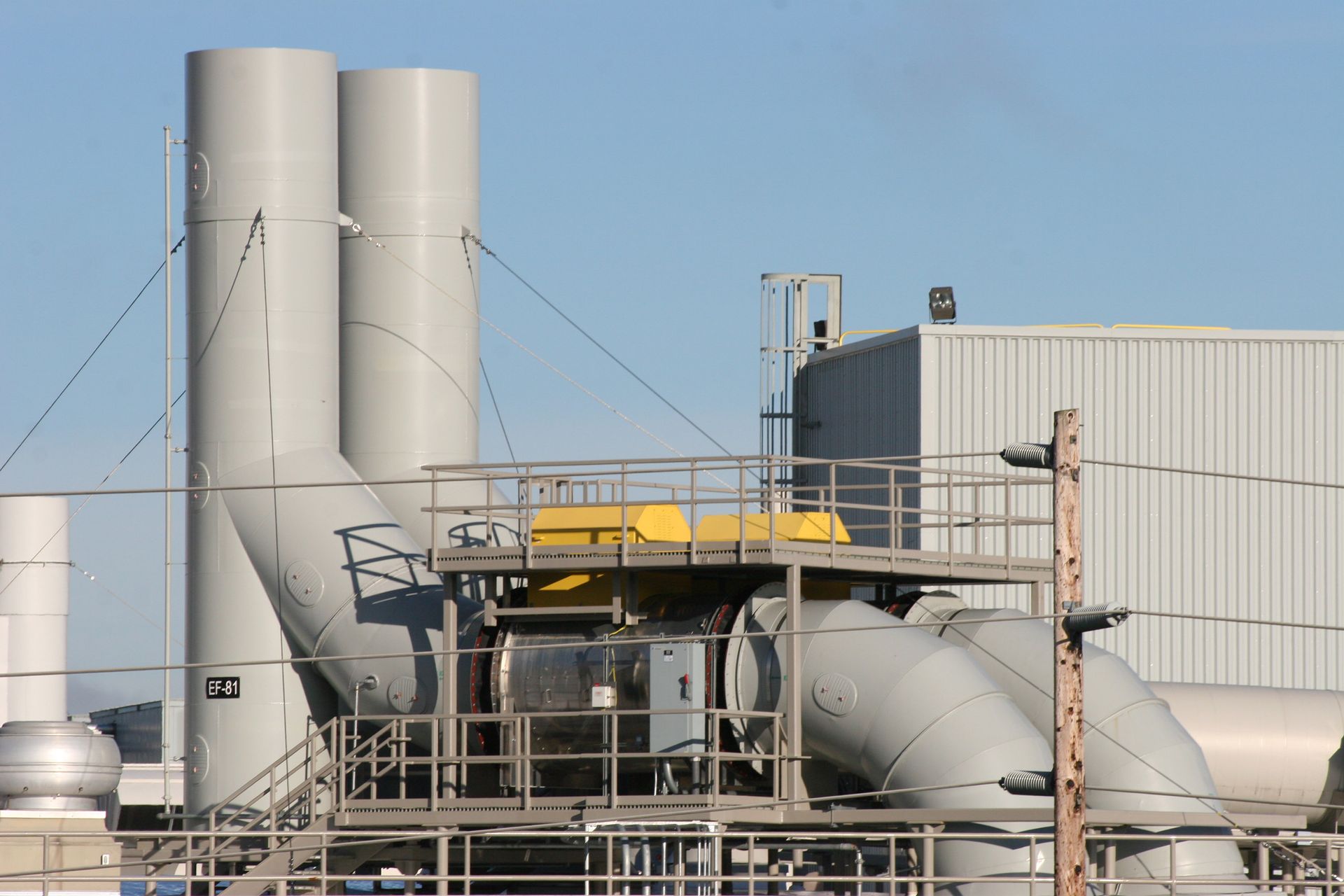 Industrial facility with grey smokestacks, pipes, and metal walkways against a blue sky.