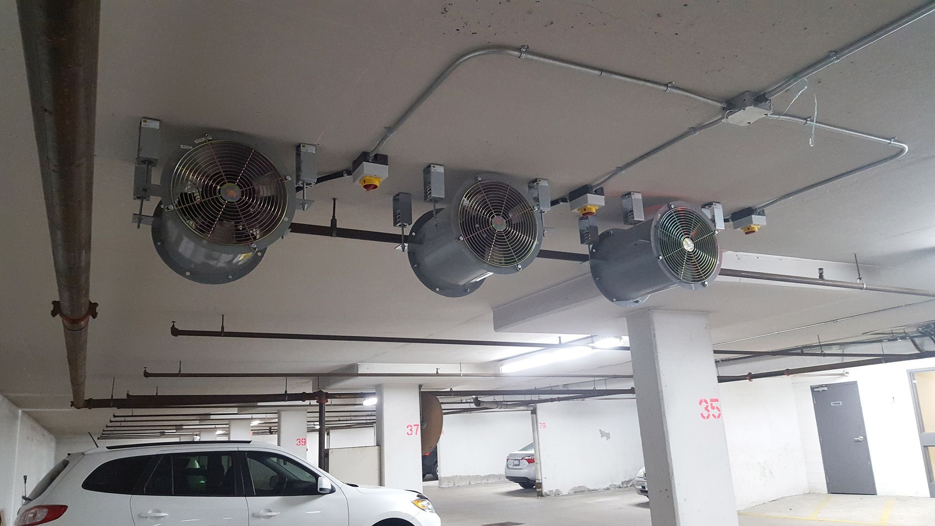 Three large ventilation fans installed on a parking garage ceiling, with a white car visible below.