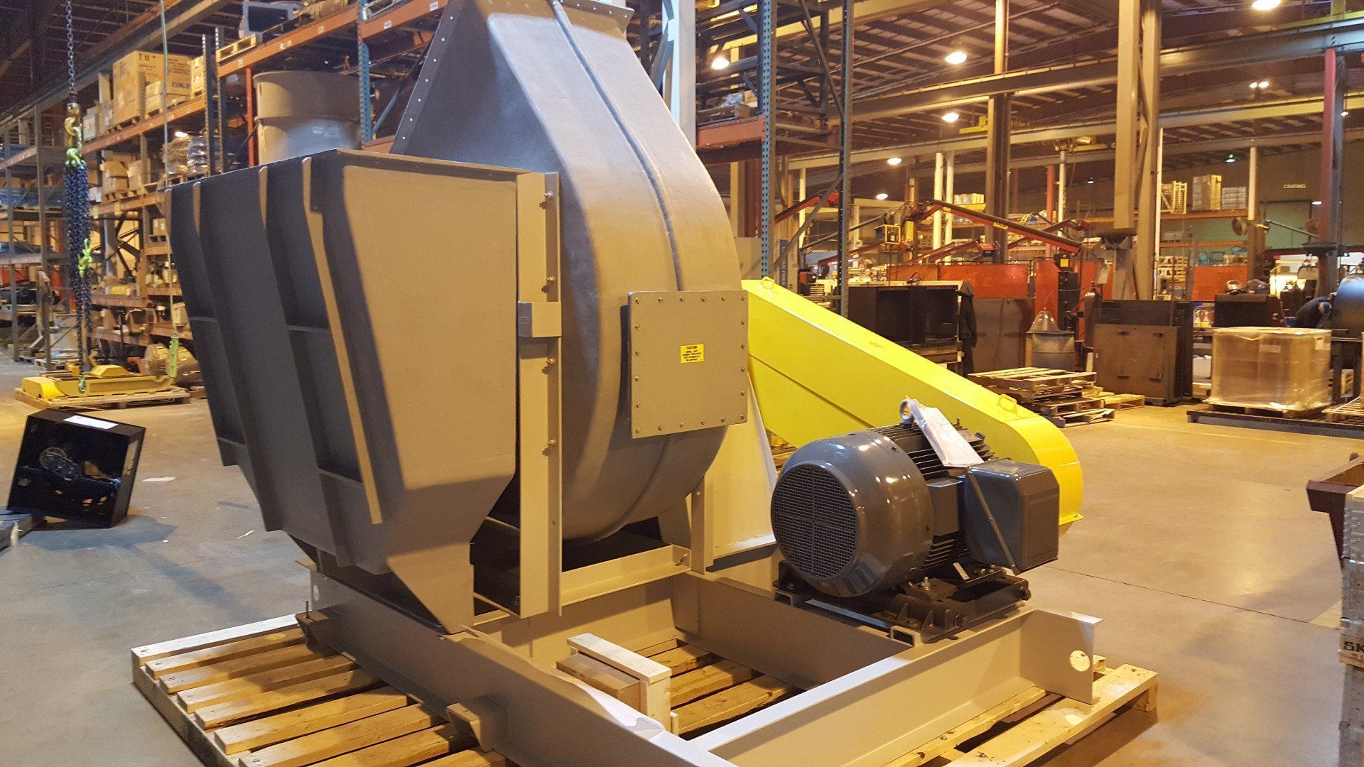 Large industrial fan, gray with yellow belt guard, mounted on a pallet in a warehouse.