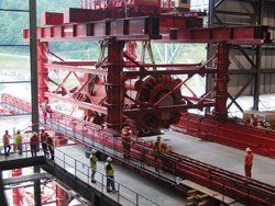 Red industrial crane lifting a large, round mechanical component indoors, with workers observing.