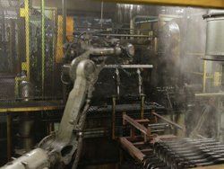 Industrial robot arm placing metal parts into a furnace in a factory setting. Steam visible.