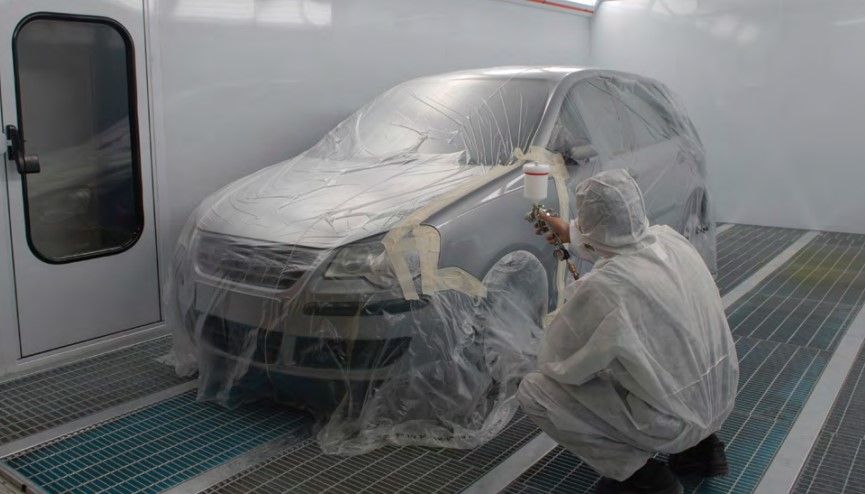 Person in protective suit spraying paint on car inside a paint booth. Car is partially covered in plastic.