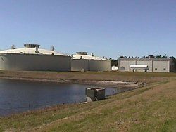 Large industrial tanks and a building next to a pond under a blue sky.
