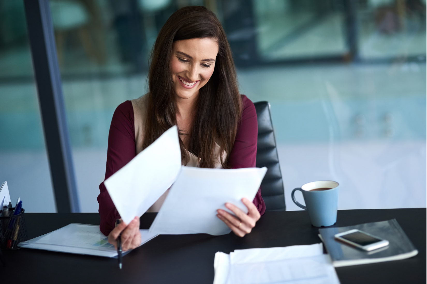 Woman smiles, reviews papers at desk. Coffee cup, phone, pen holder, and window visible.