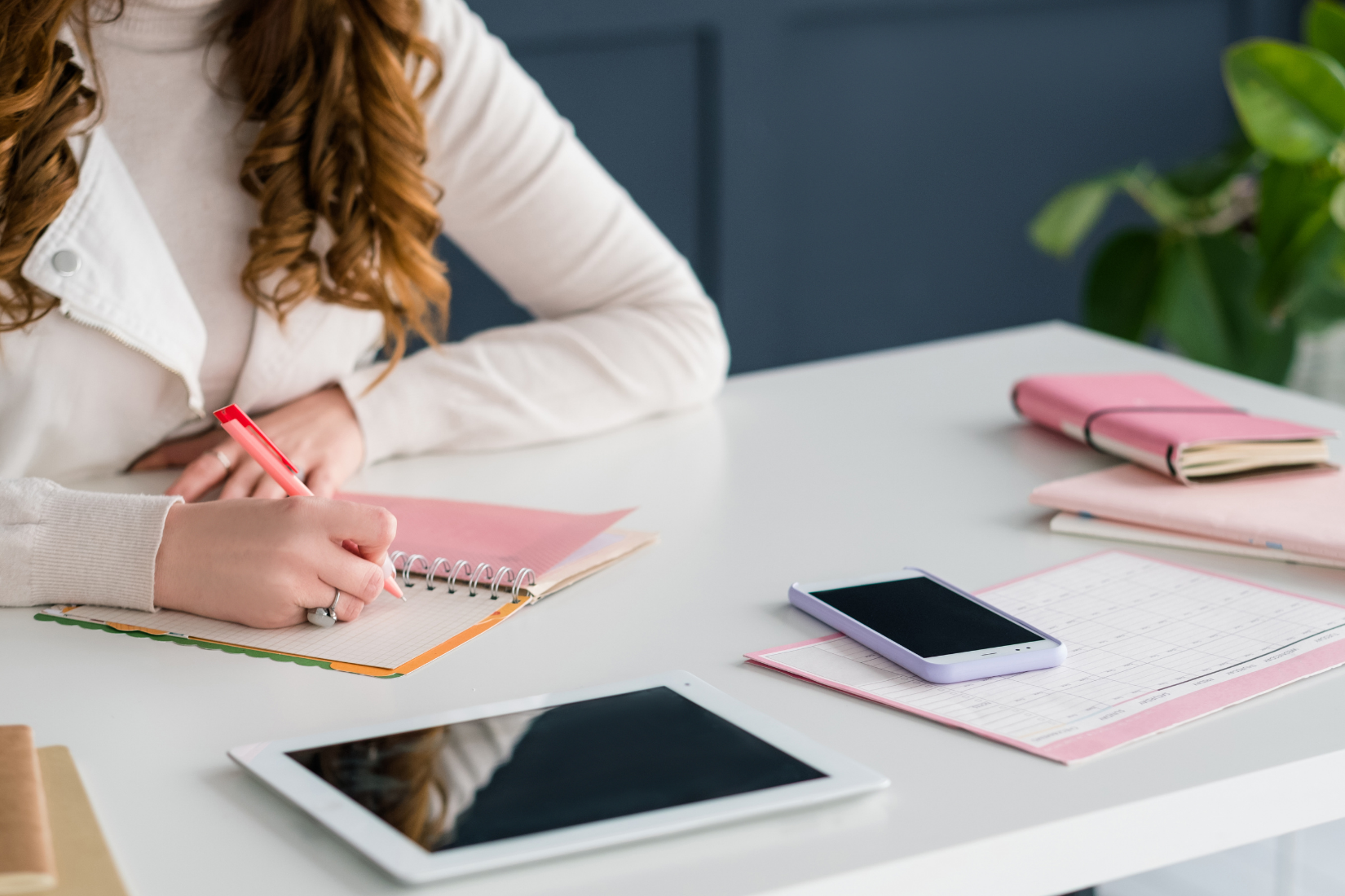 Woman writes in a notebook at a white desk with a tablet, phone, and other pink office supplies.
