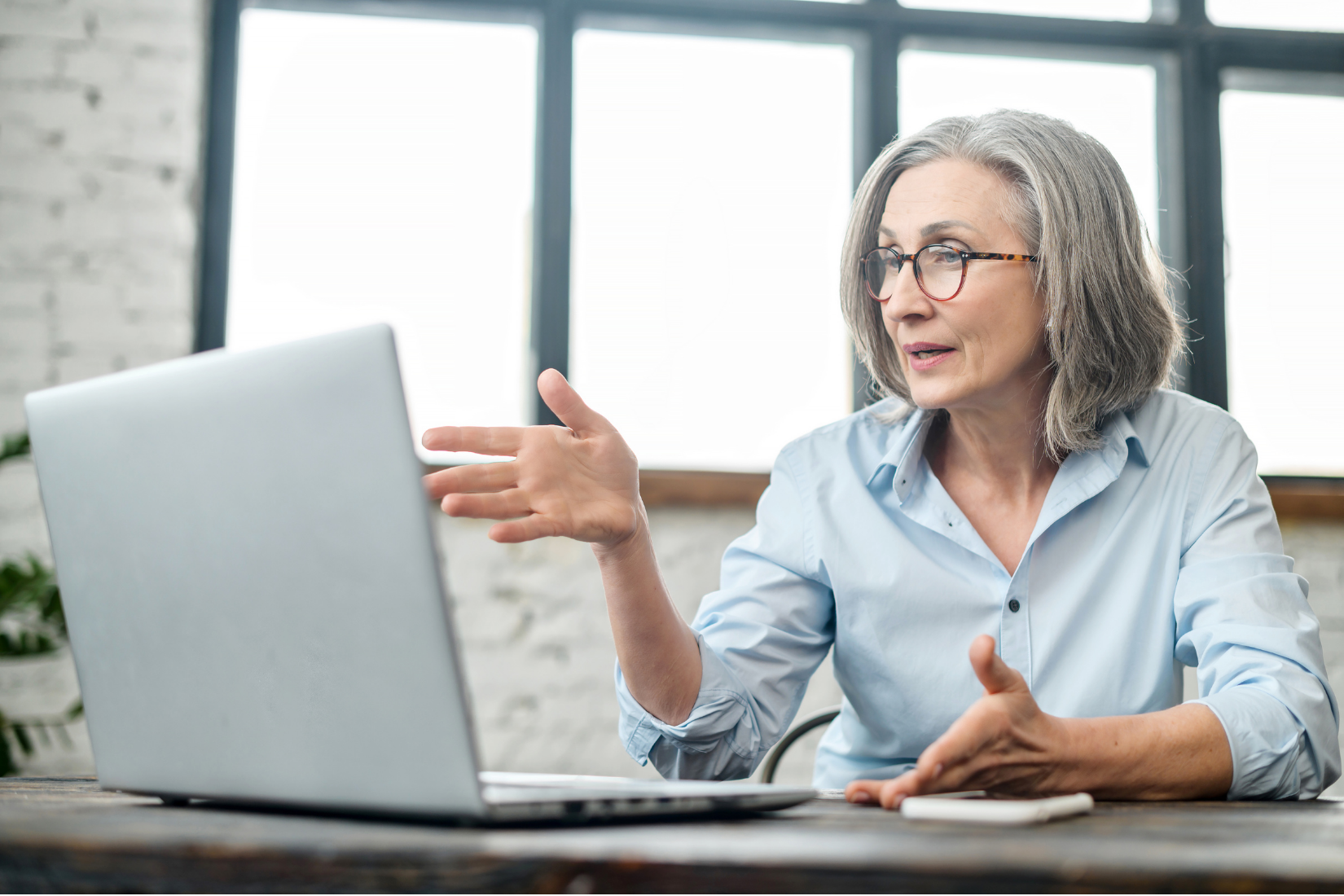 Woman speaking in a video call from a bright office, gesturing beside a laptop