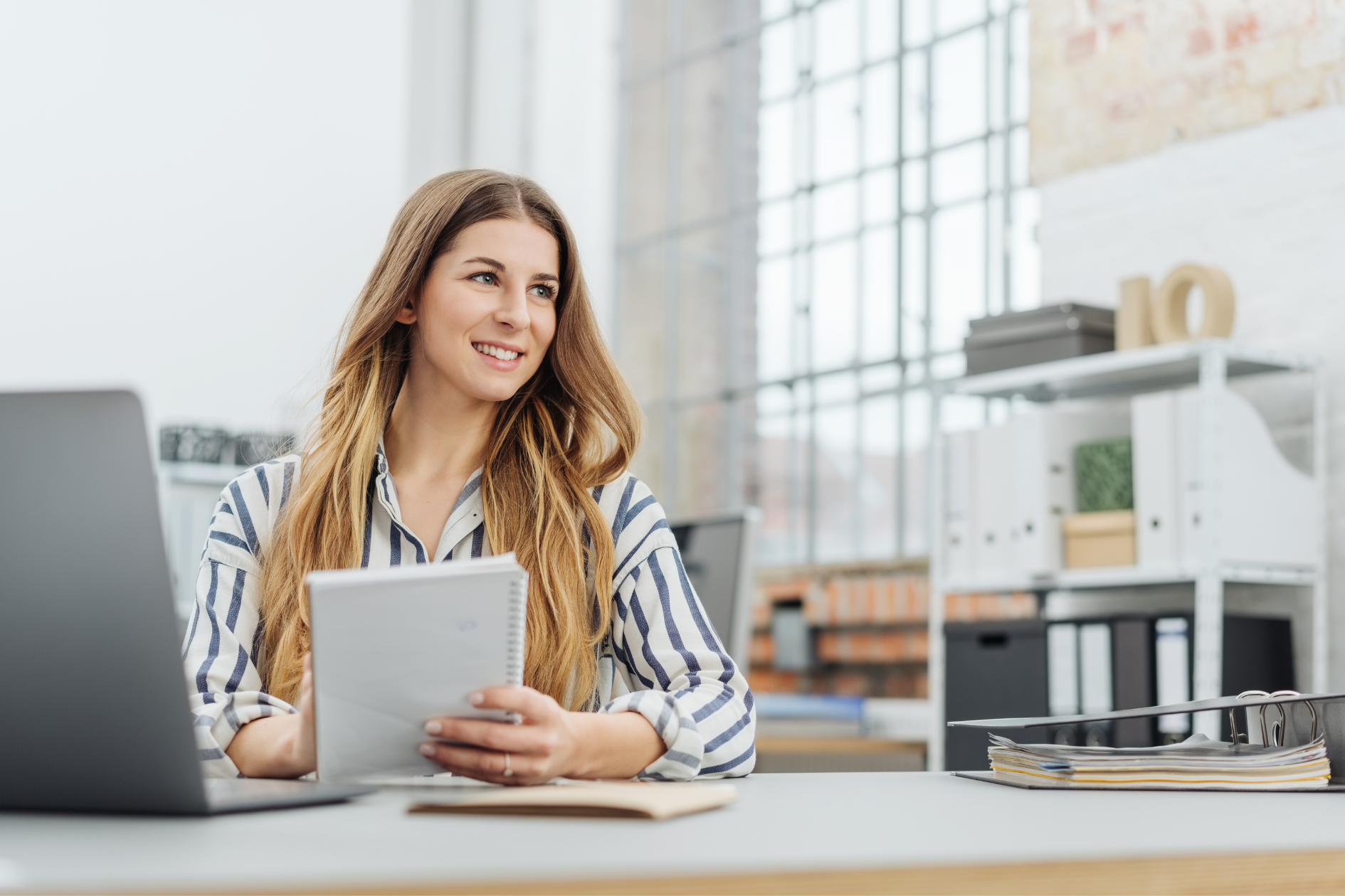 Woman on phone at desk, writing notes beside a laptop in a modern office.