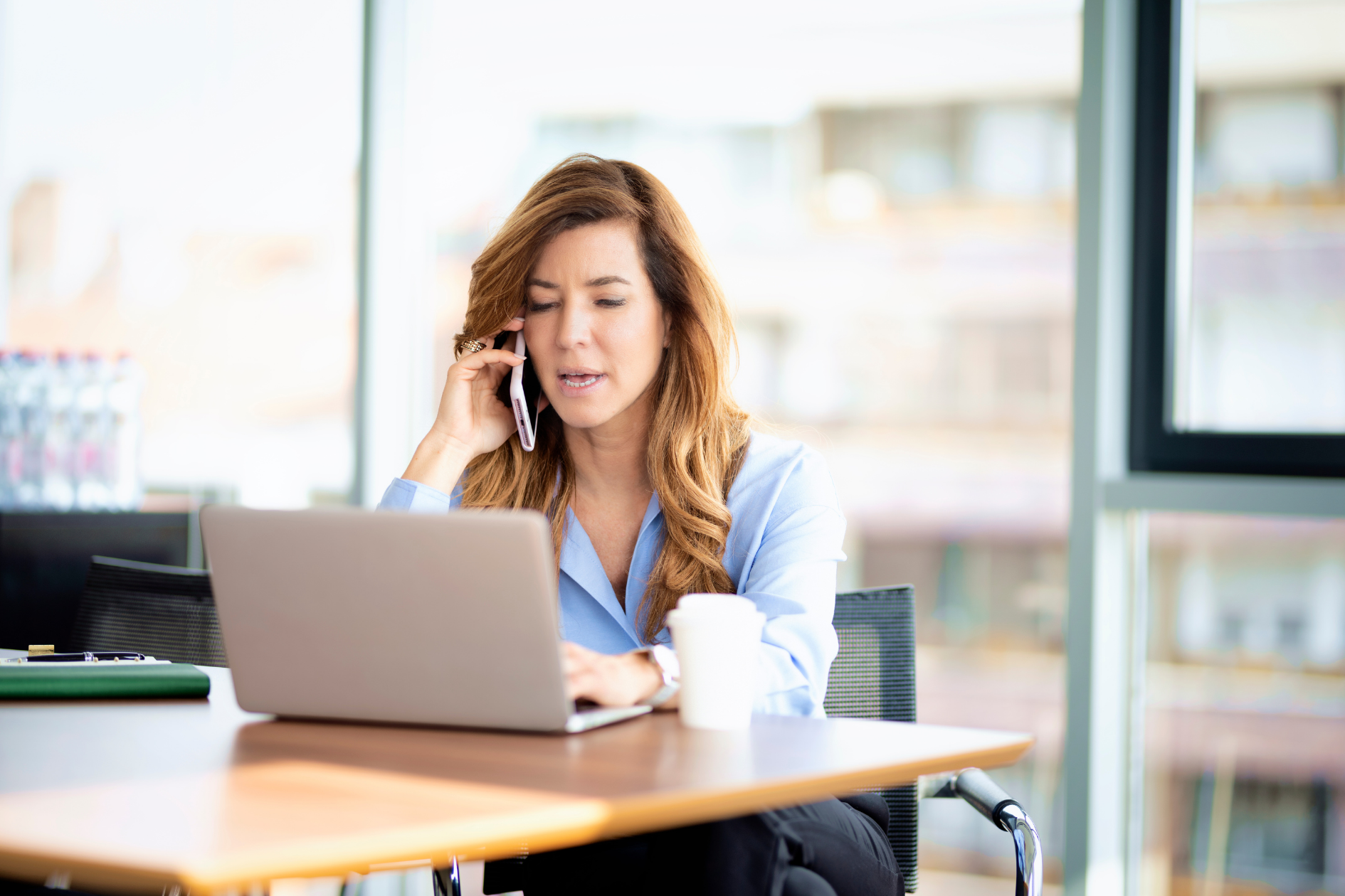 Person talking on a phone while working on a laptop at a desk in a bright office