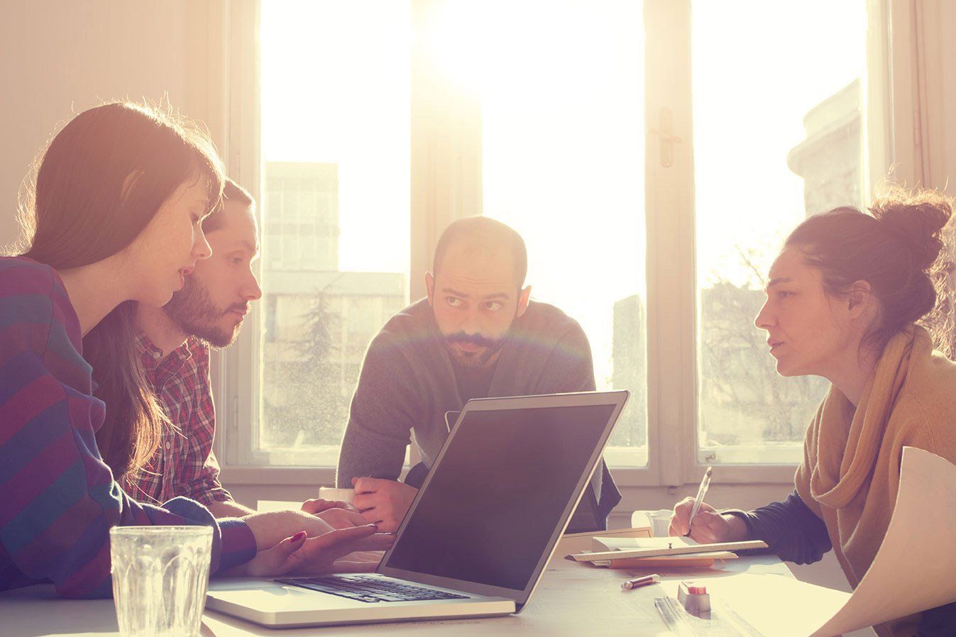 People gathered around a laptop in a sunlit meeting room, discussing together.