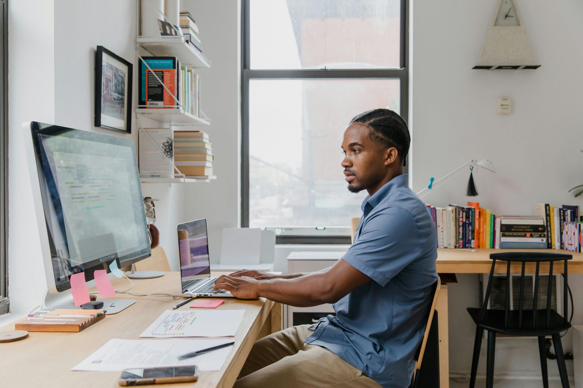 Man in blue shirt at desk, working on computer, beside a window.
