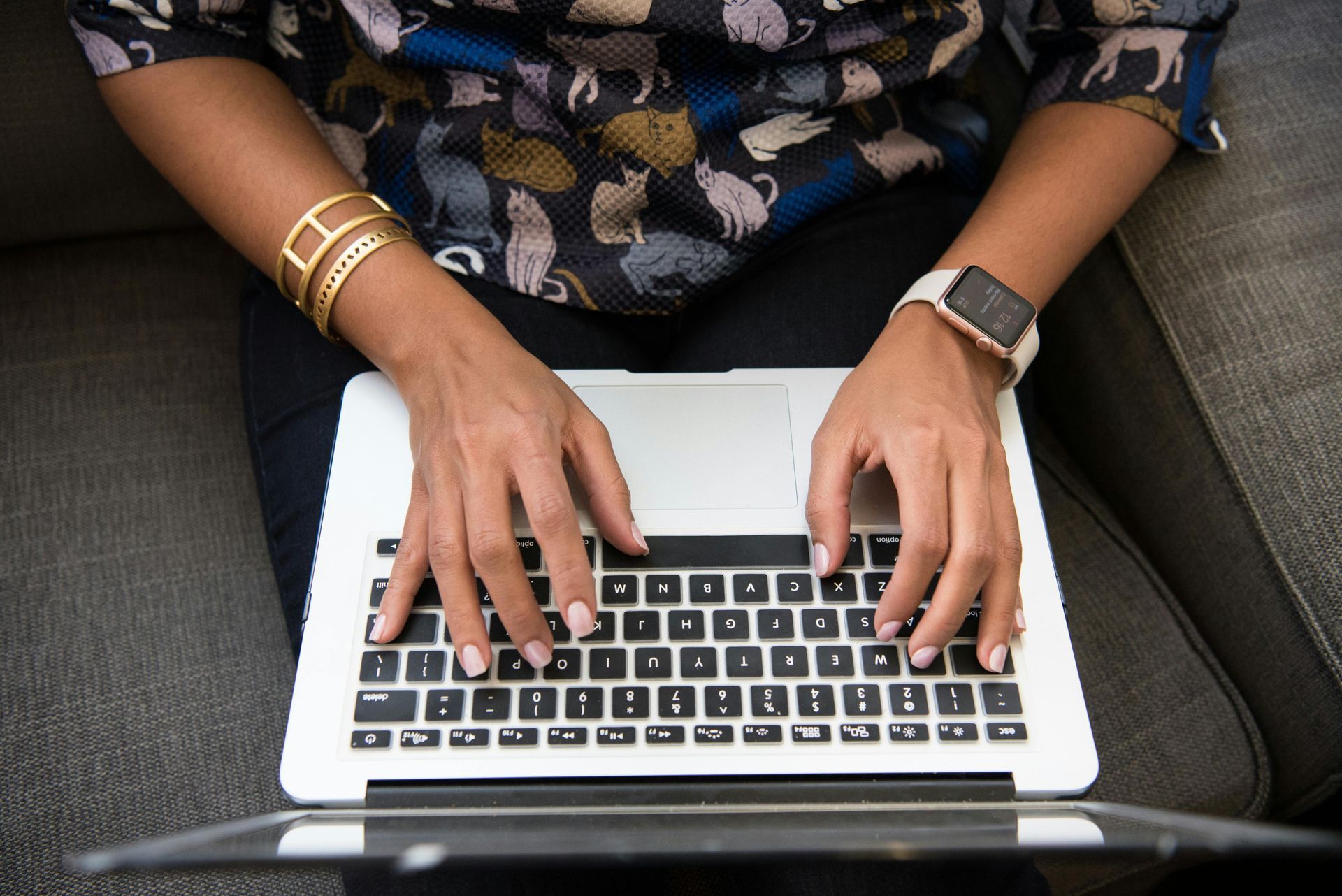 Person typing on a laptop while seated on a gray couch, wearing a patterned shirt and smartwatch.