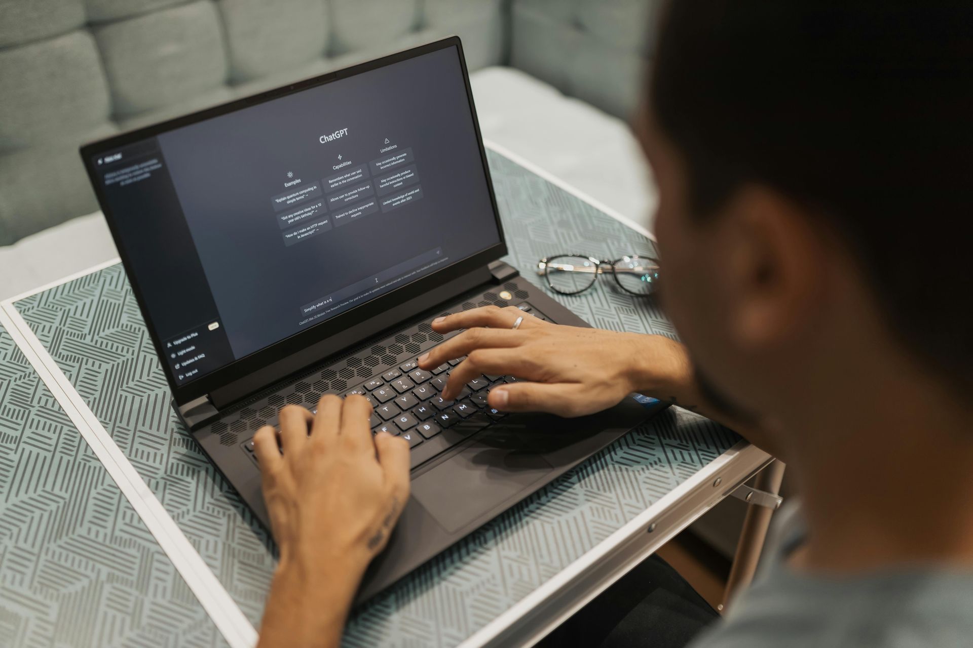 Person typing on a laptop, with eyeglasses on the side. The laptop displays a dark interface with several boxes.