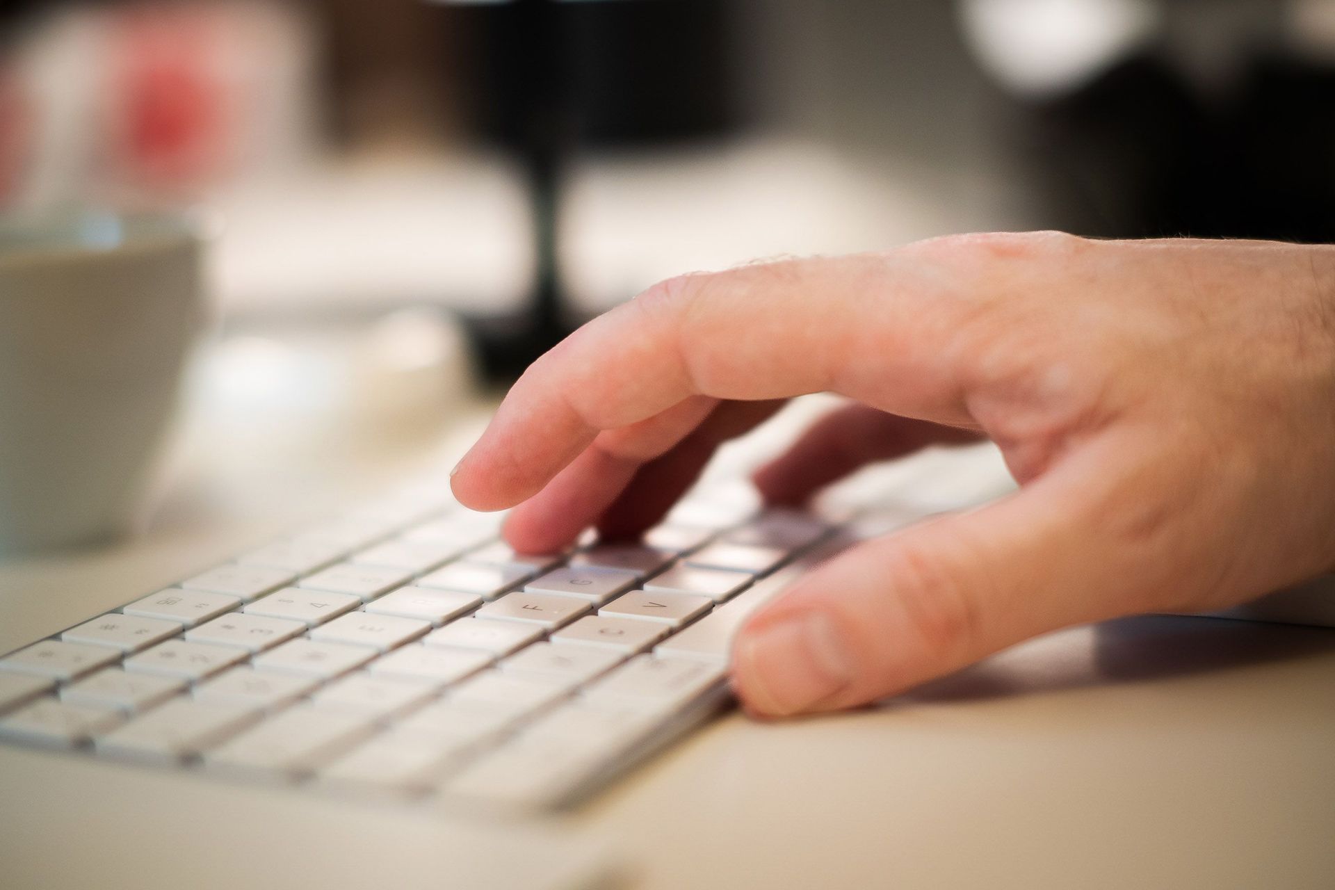 Hand typing on a white keyboard beside a blurred coffee cup