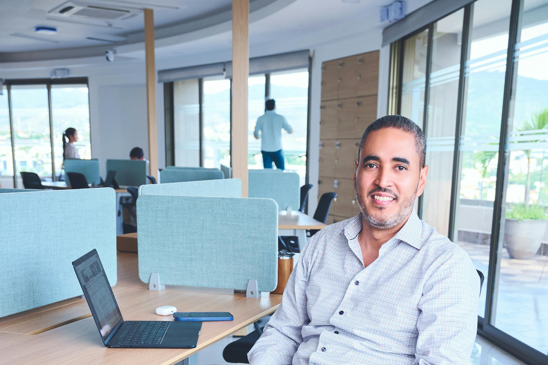 Smiling man seated in a bright office with a laptop and large windows in the background