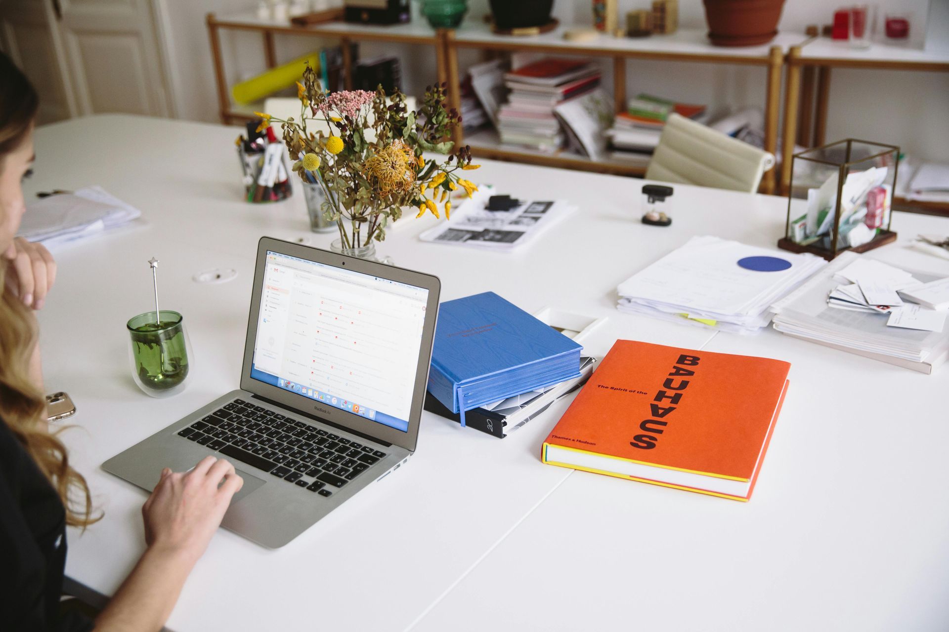 Woman working on a laptop at a white desk; books, flowers, and supplies are nearby.