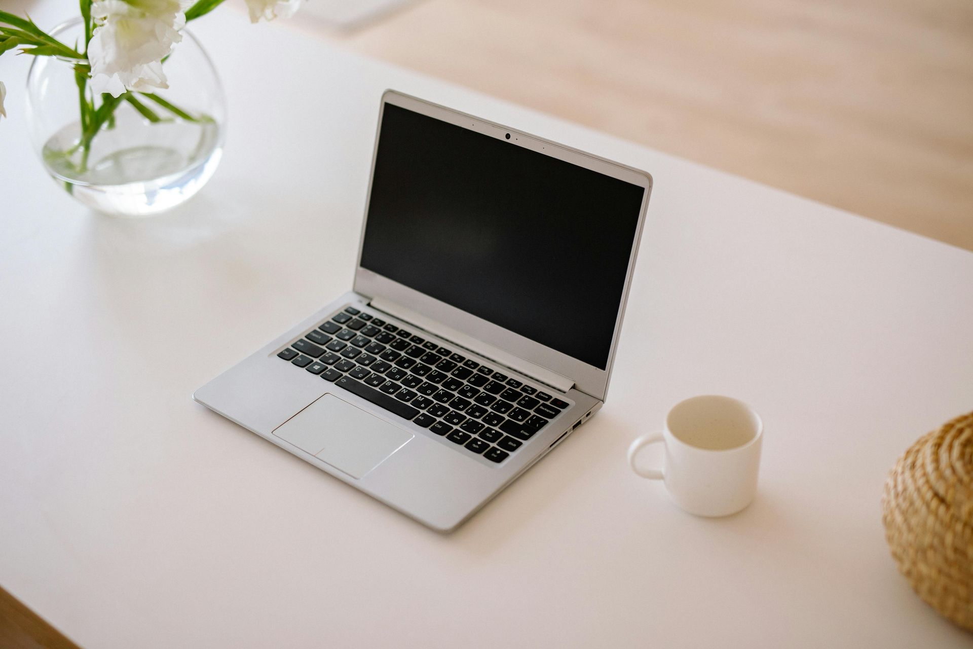 Minimal desk setup with an open laptop, white mug, small plant, and woven decor on a light table.