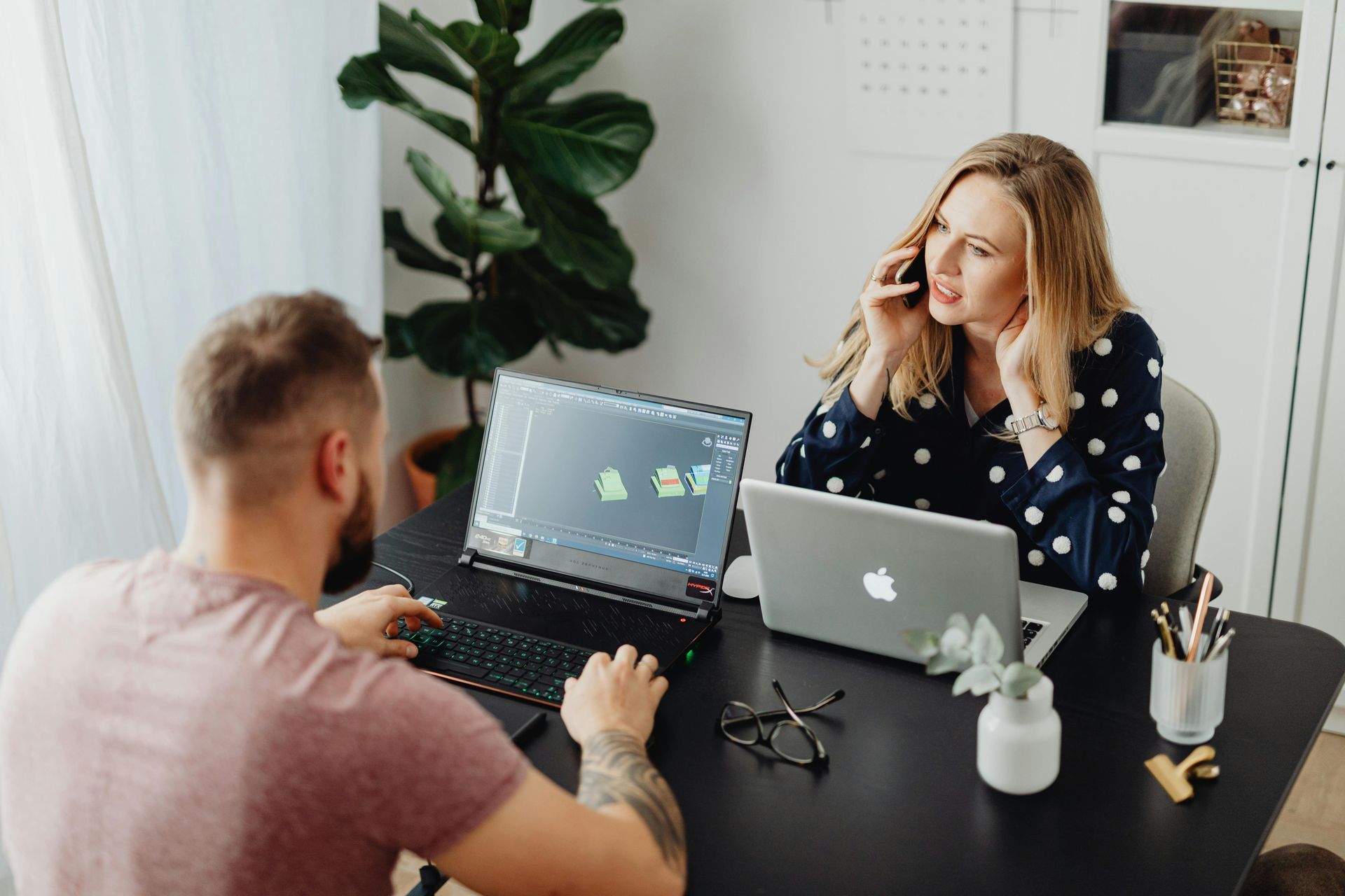 Woman on phone, man working at computers, in bright office setting.