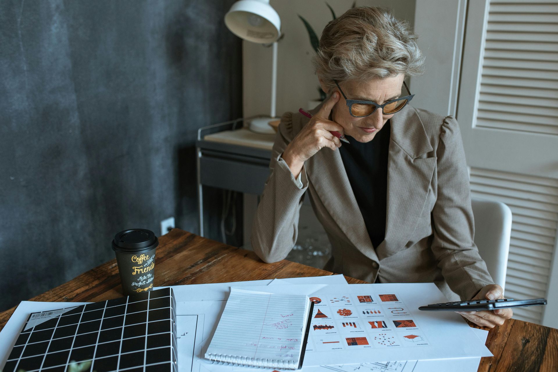 Woman in glasses, blazer, studying drawings at a wooden desk with a coffee cup.