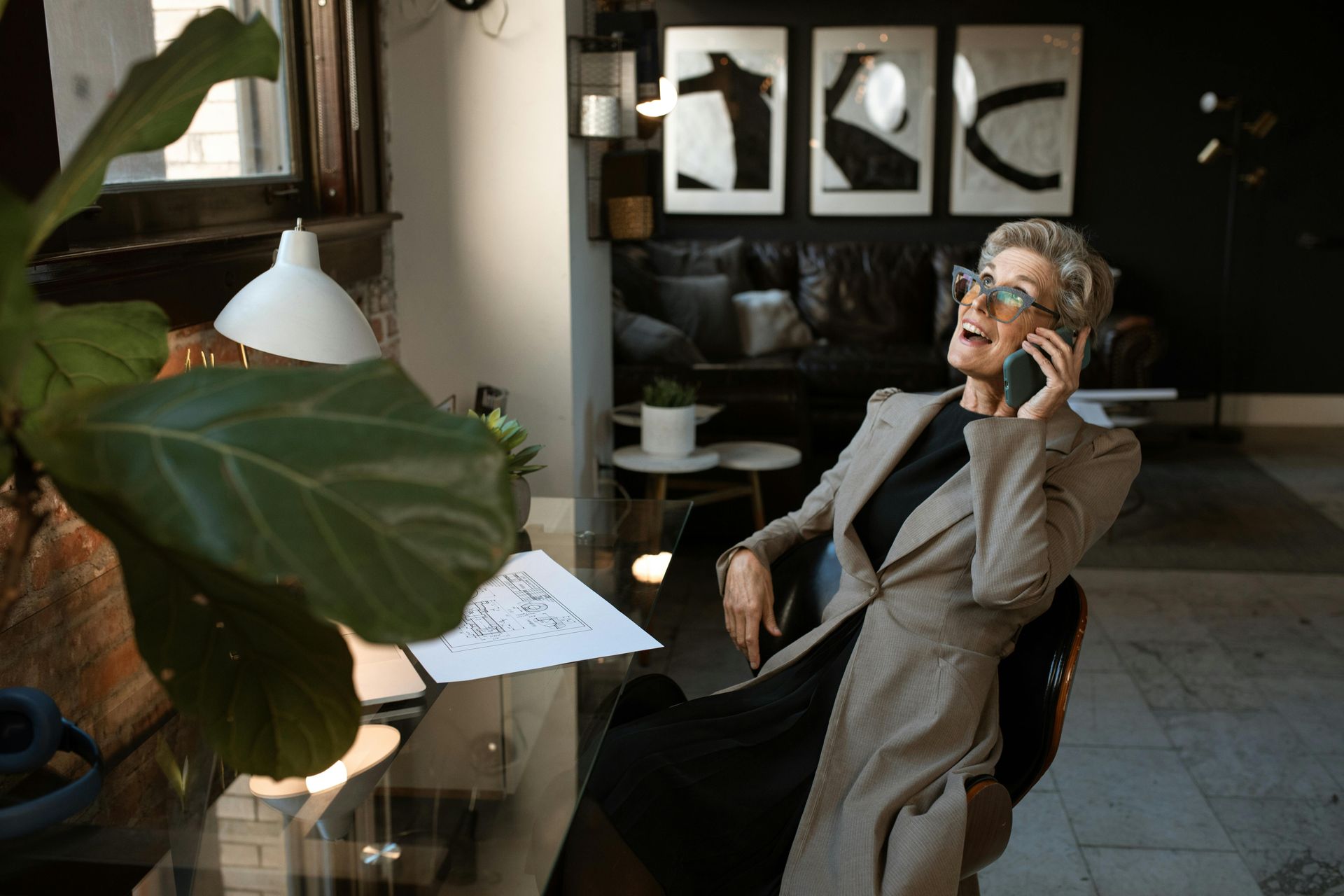 Woman in coat laughs on phone at a desk in a stylish room with art and a plant.