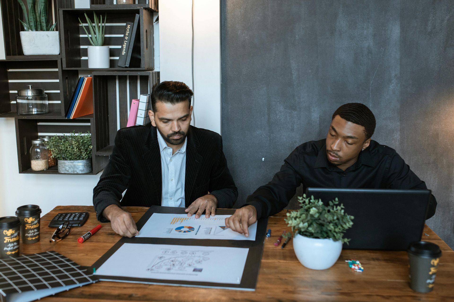 Two men, one in a blazer, reviewing documents at a wooden desk. A laptop, plants, and office supplies are present.