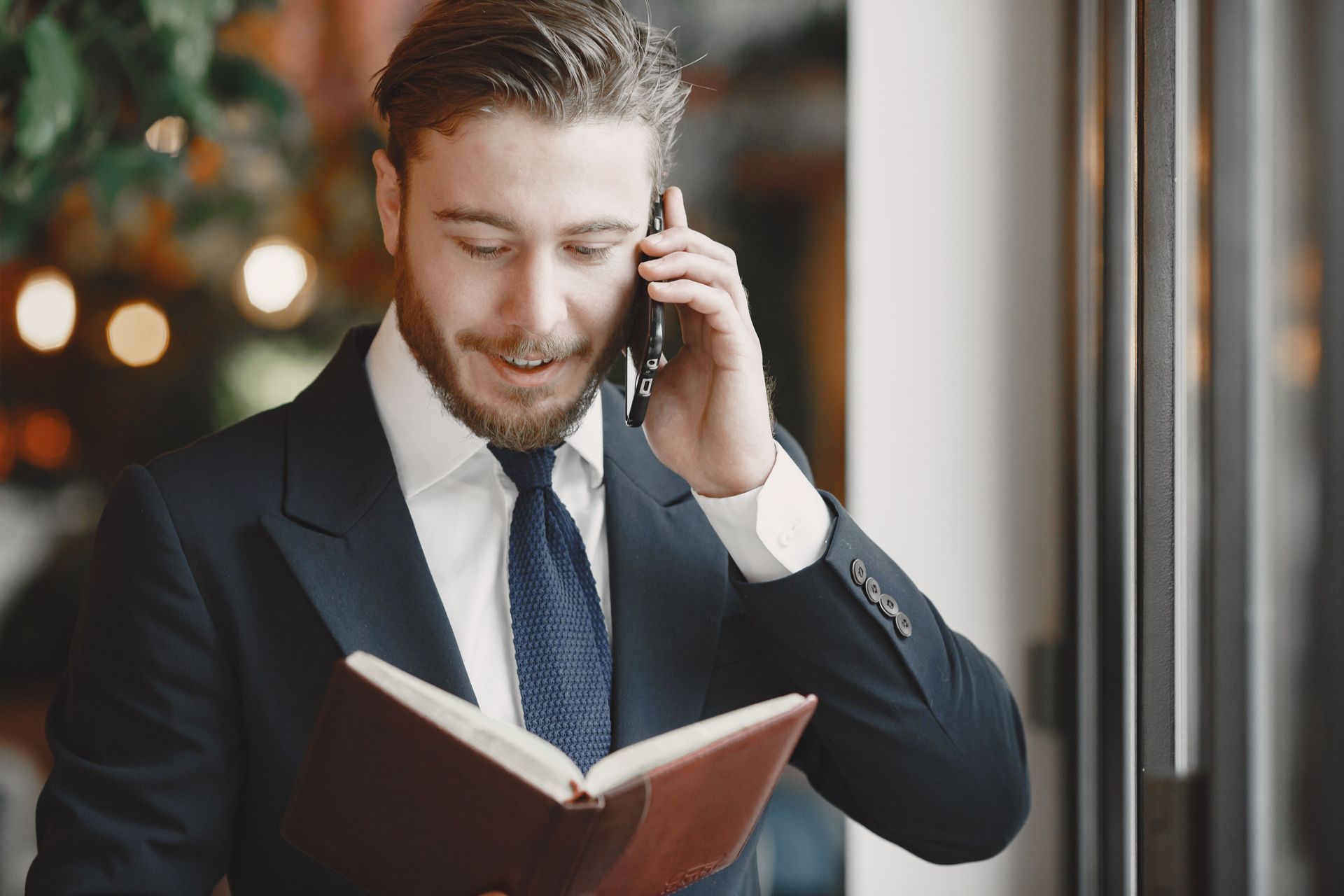 Businessman in a dark suit reading a book while talking on a phone by a window