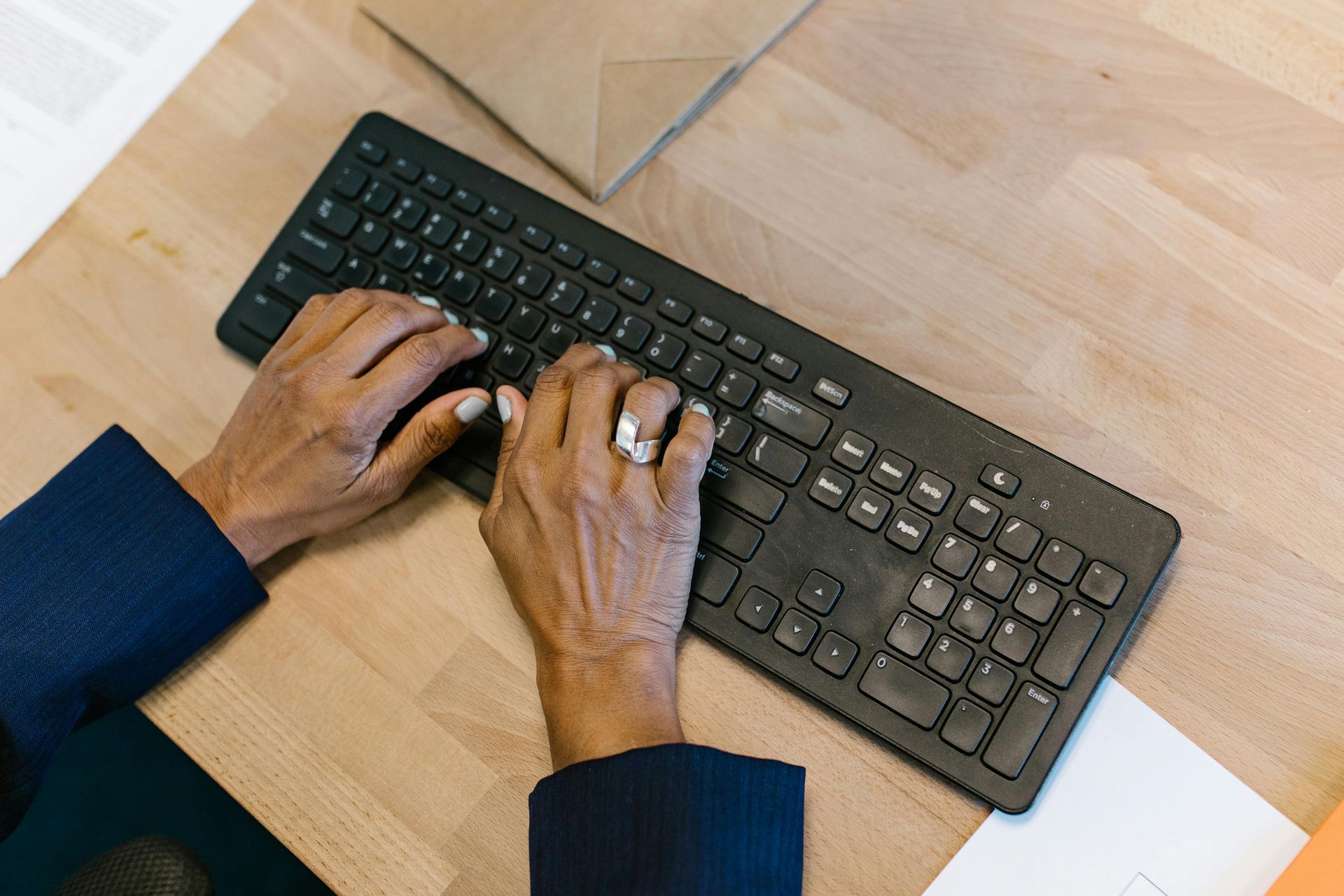 Hands typing on a black keyboard on a light wooden desk with a white notebook nearby