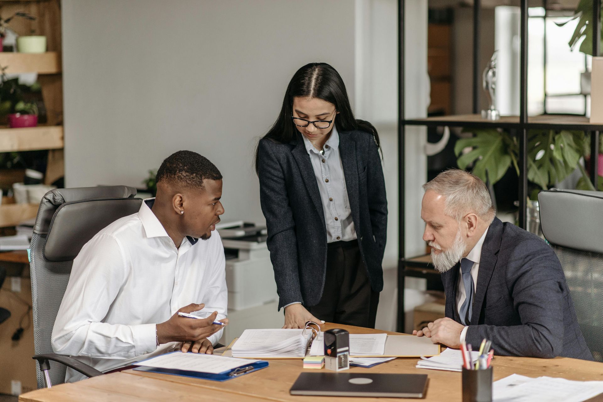 Three office workers reviewing documents at a desk. One woman stands, looking down. Two men seated, gesturing.