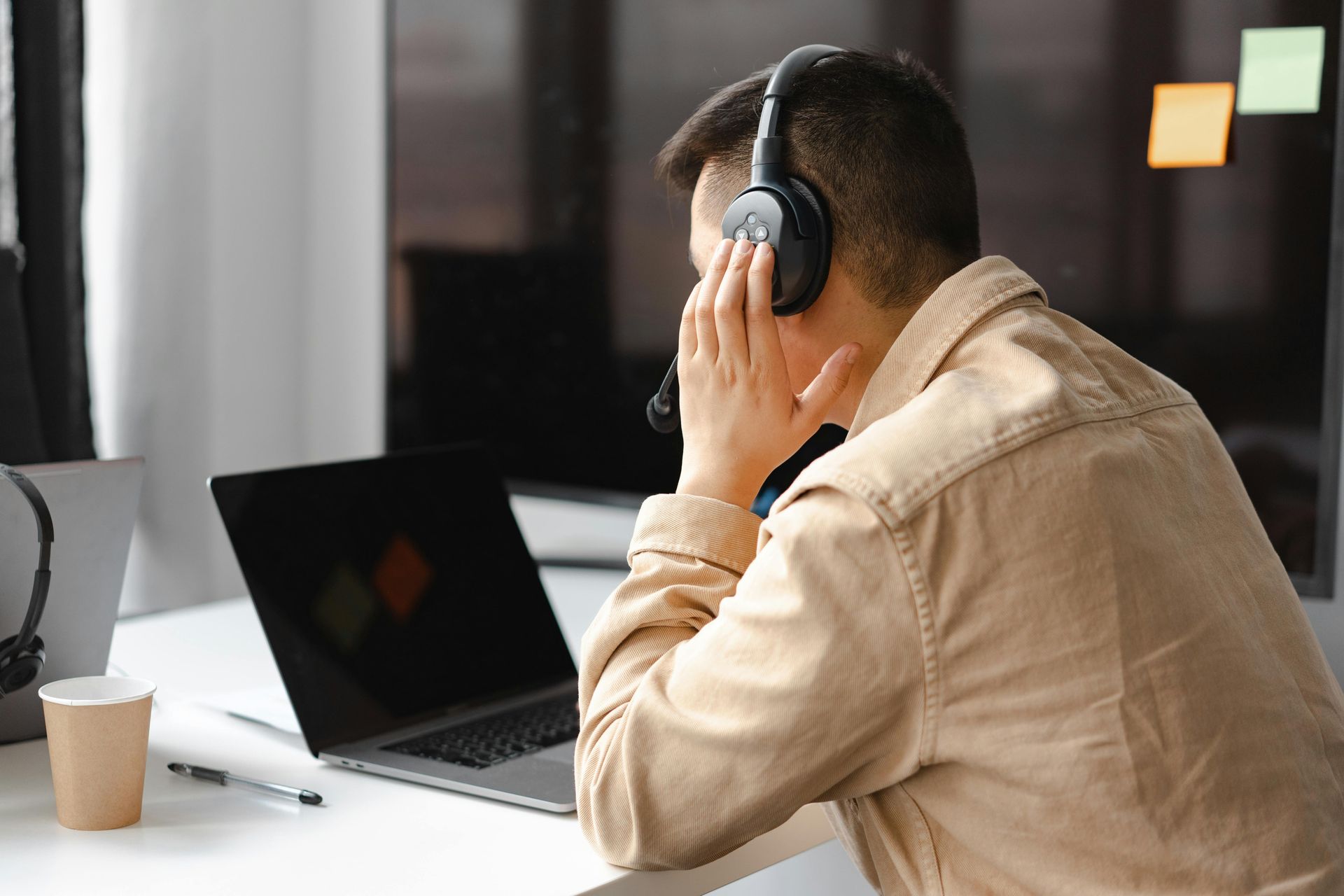 Man wearing headset, hands on face, at laptop. Beige shirt, white desk, indoor office.