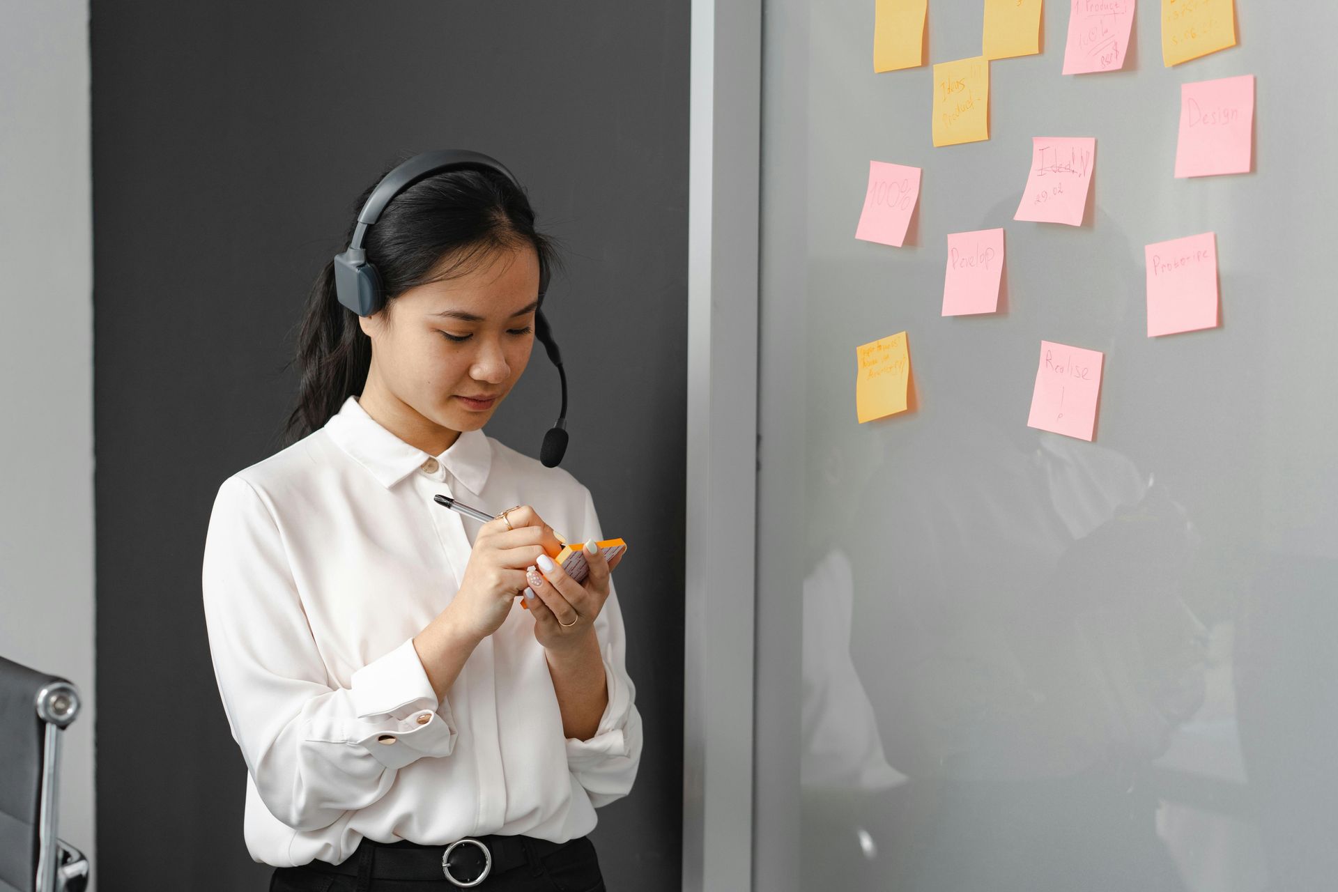 Woman with headset, writing on a notepad in front of a wall with sticky notes.